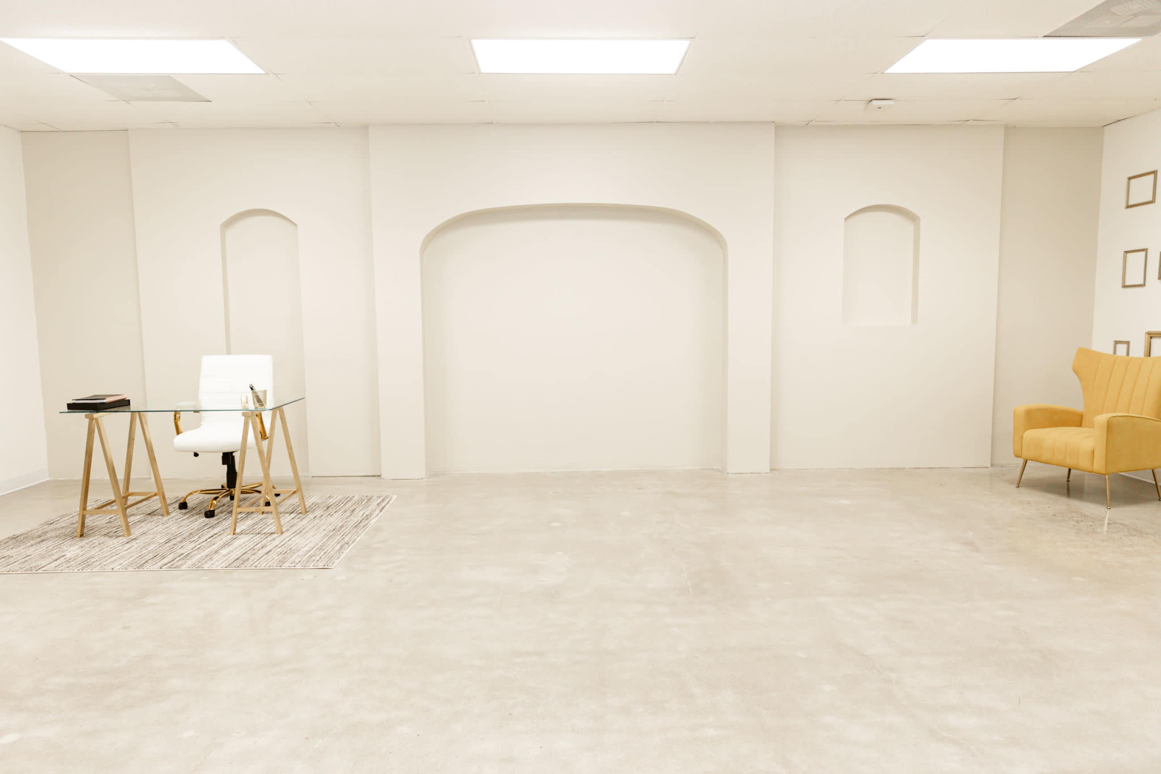 A minimally furnished office space with a glass desk, a white chair, and a yellow armchair against a plain wall.