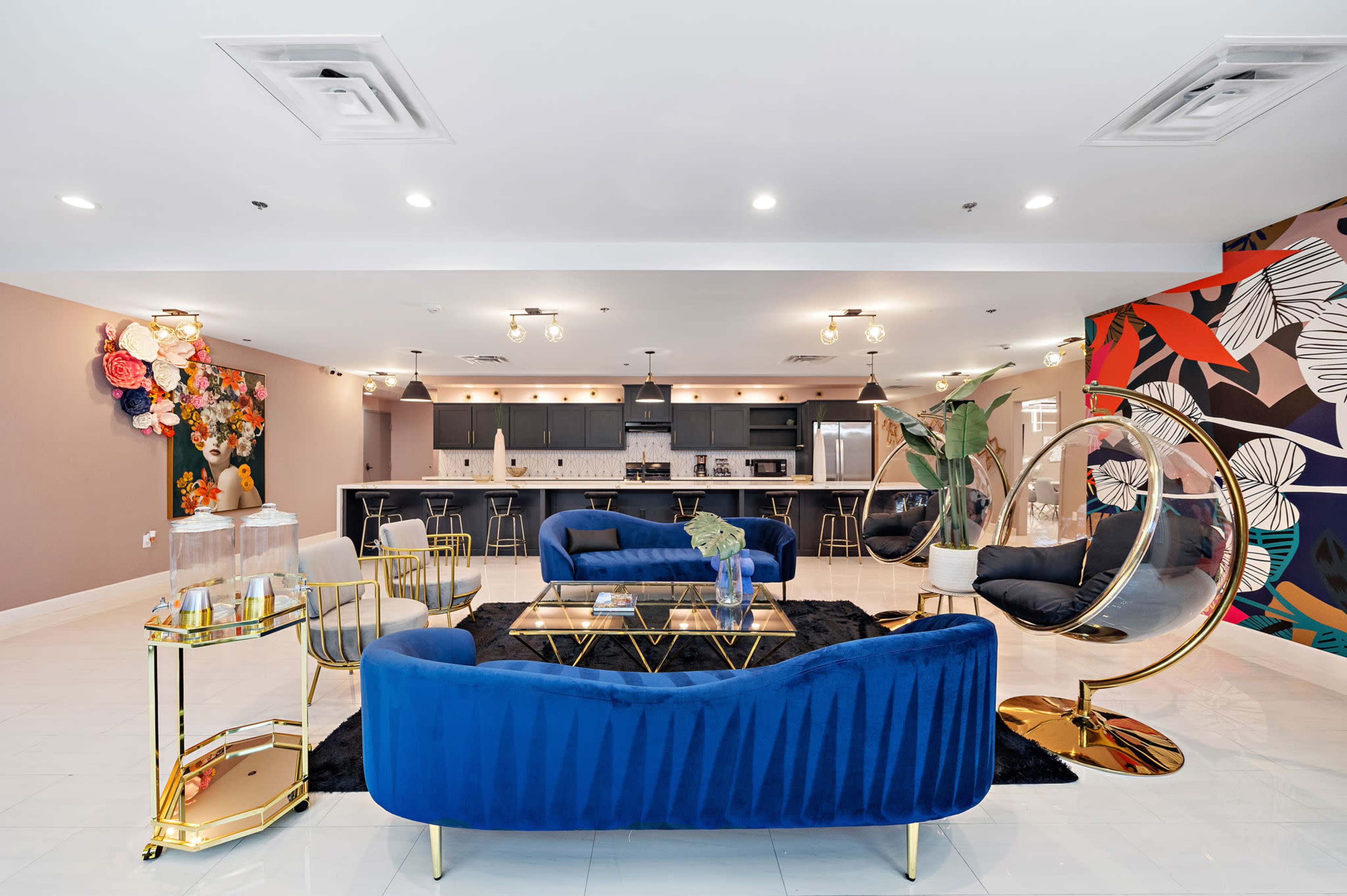A modern lounge area with blue velvet seating, gold accents, and a colorful accent wall featuring floral designs, leading to a kitchen space in the background.