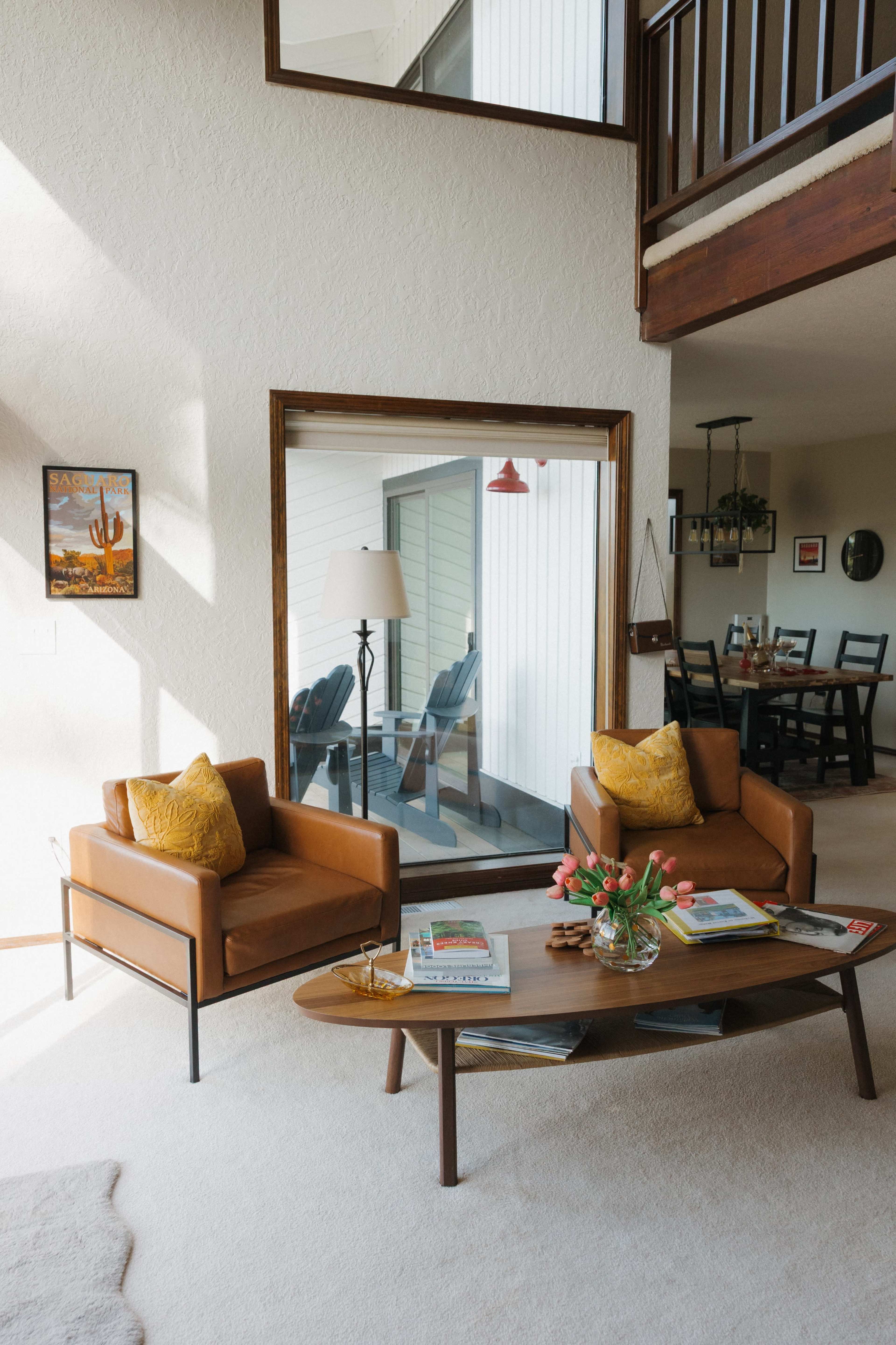 The image shows a bright living room featuring two brown chairs with yellow cushions, a round wooden coffee table with magazines and flowers, and a large window reflecting an outdoor area.