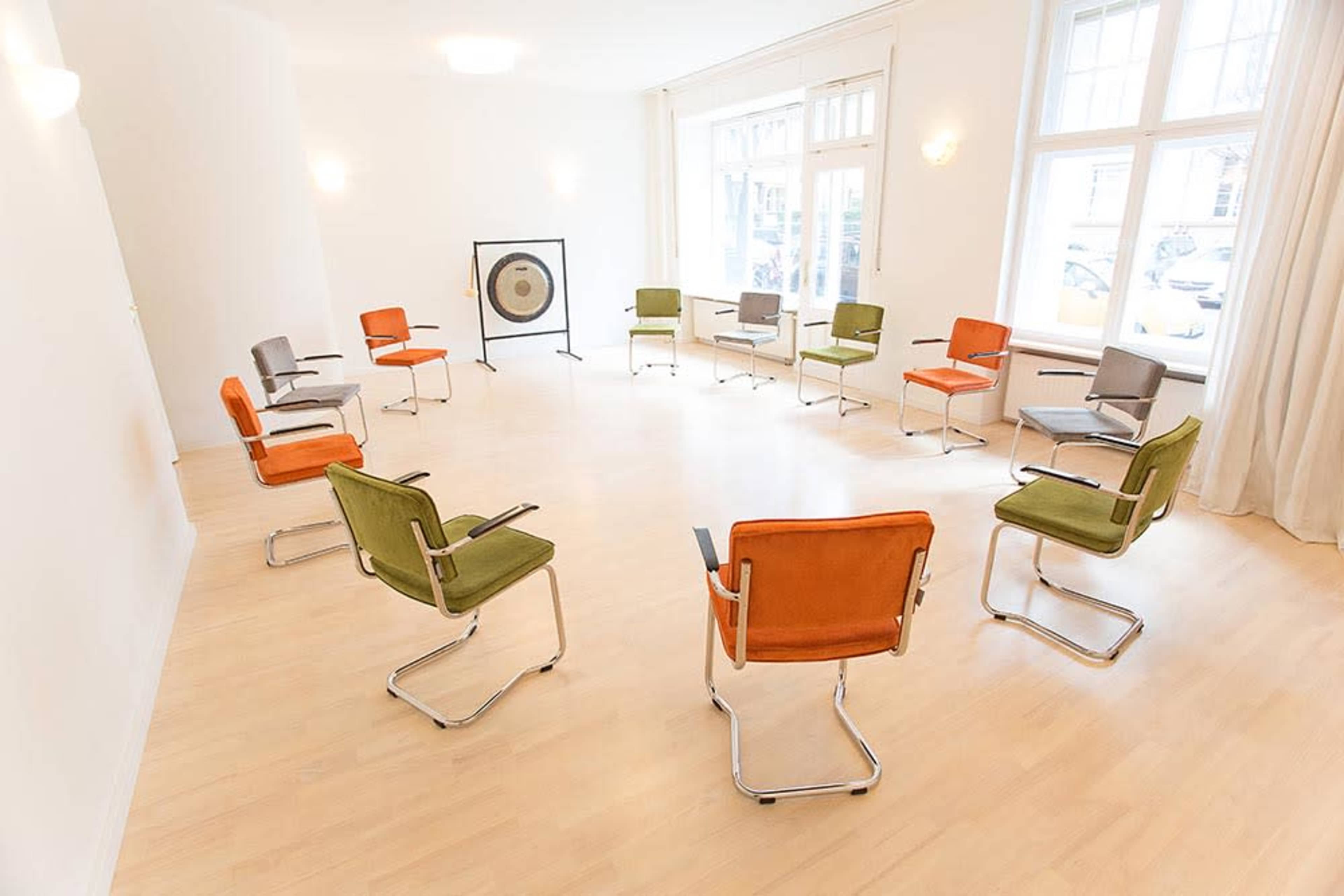 A room with a circular arrangement of eight chairs in various colors surrounding a large gong.