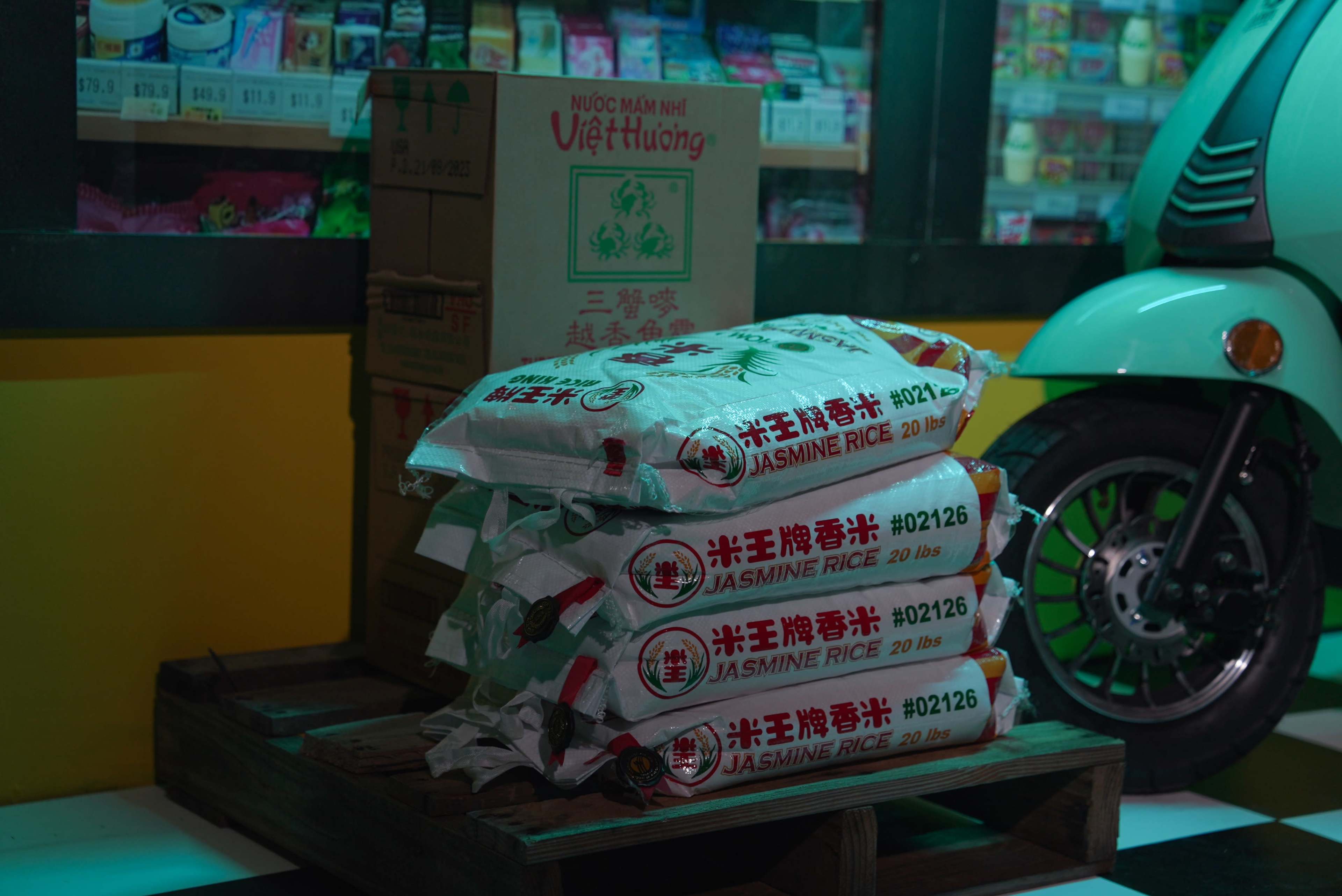 Bags of jasmine rice are stacked on a wooden pallet in front of a brightly lit store window filled with various products.