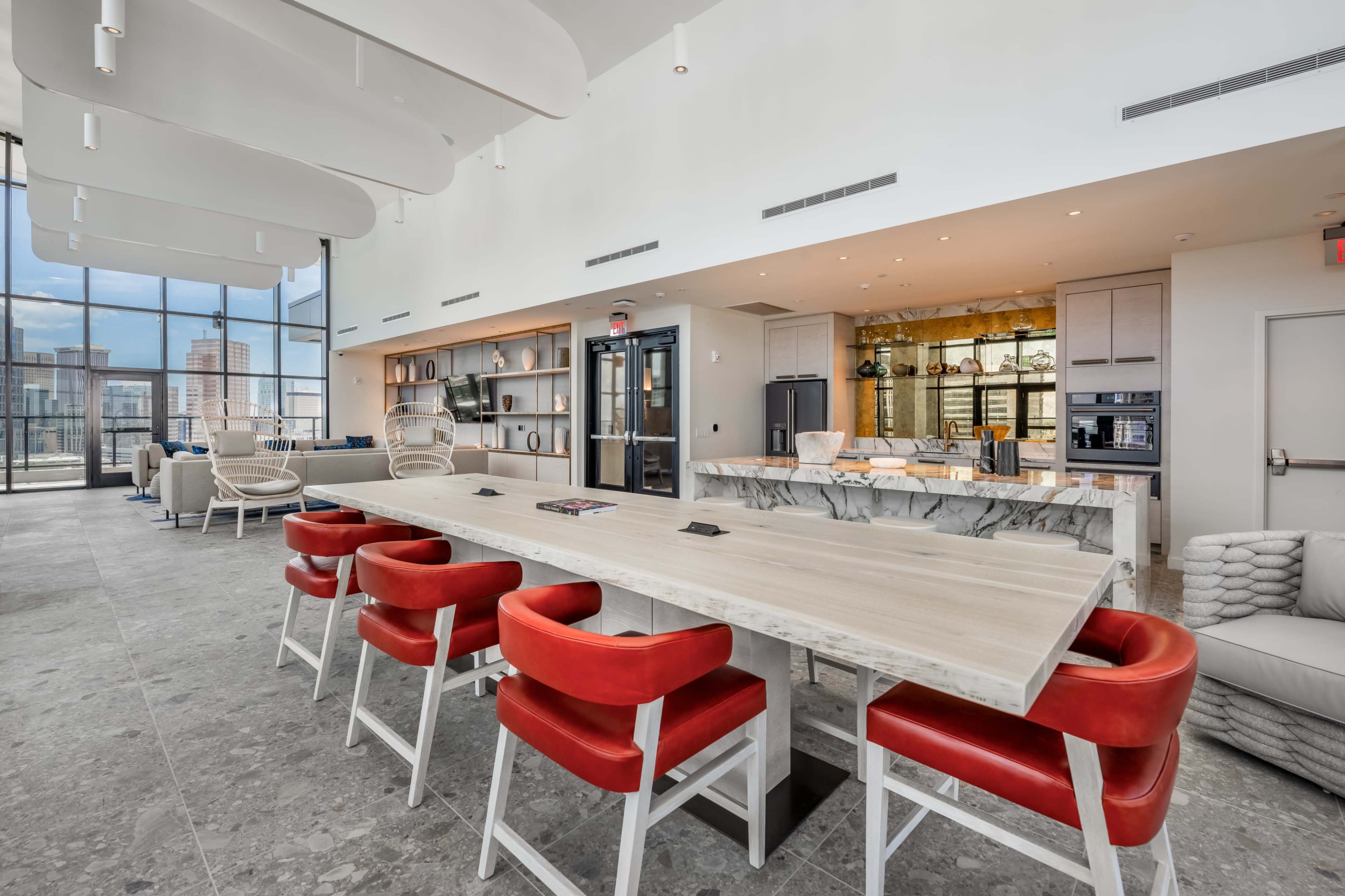A spacious modern kitchen and dining area features a long table with red chairs, large windows providing natural light, and a stylish marble countertop.