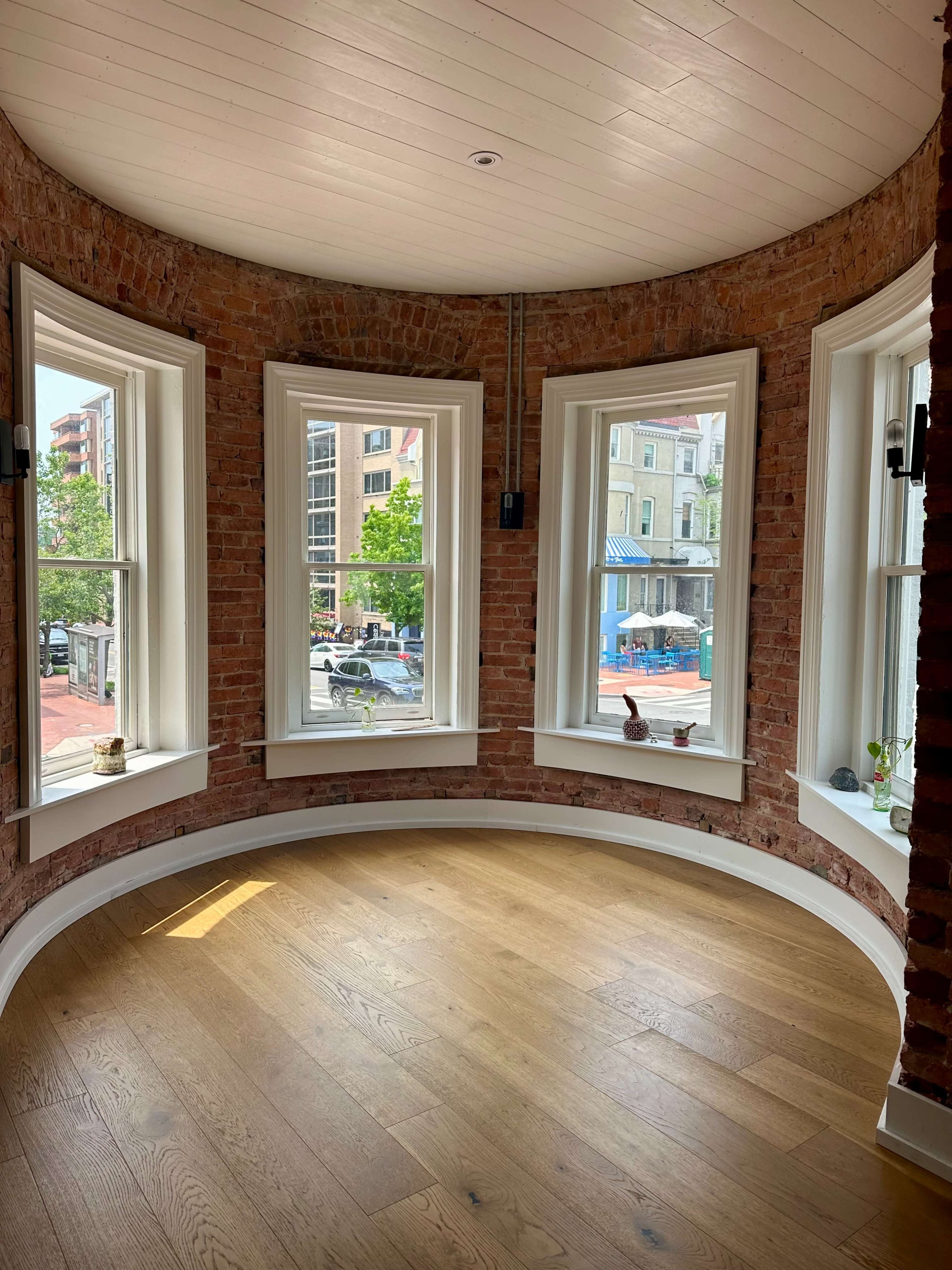 The image shows a sunlit room with large curved windows framed in white, exposing brick walls and a wooden floor.