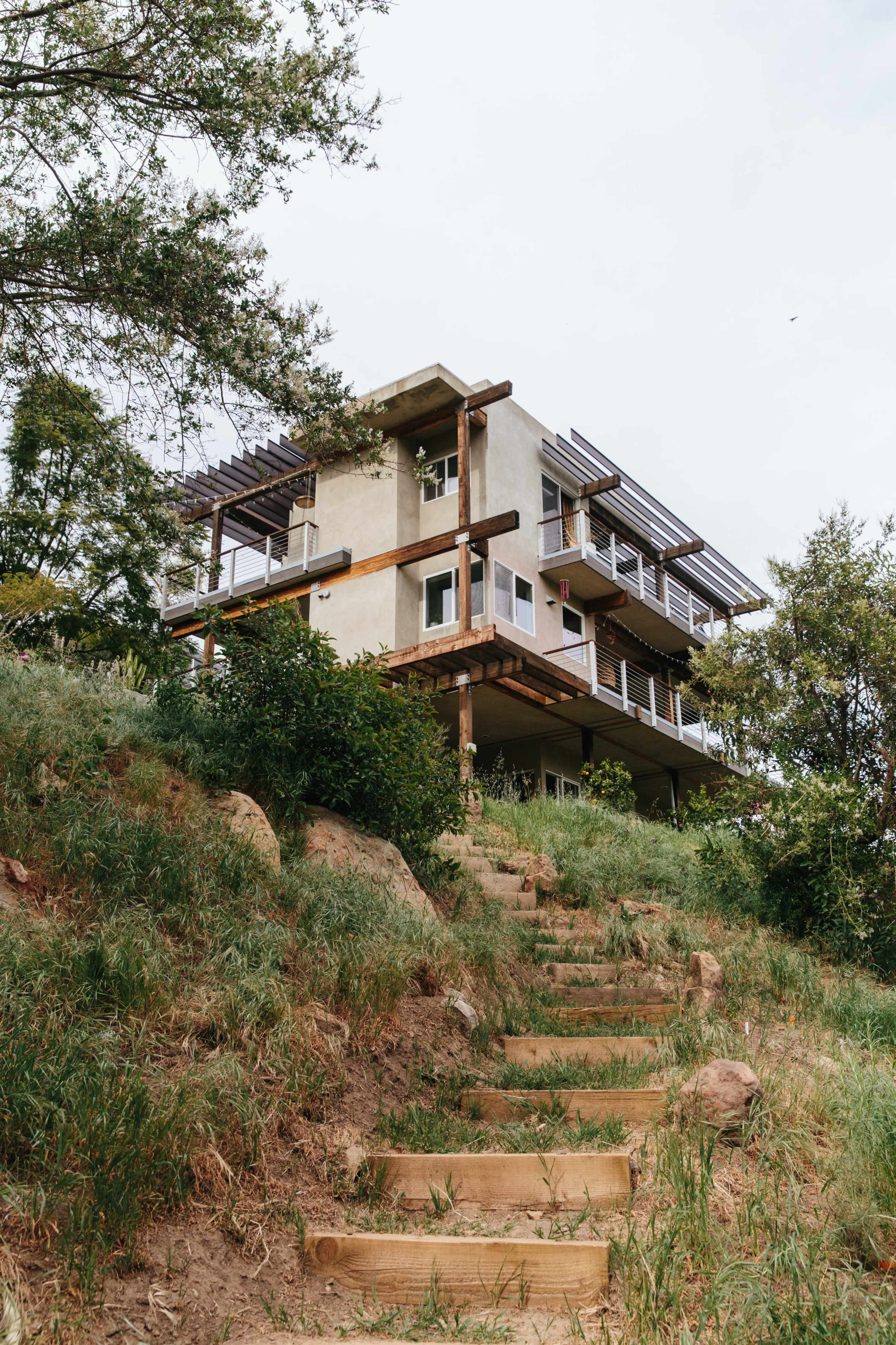 A modern house with multiple levels and a balcony is situated on a hillside, accessible by a path lined with grass and steps.