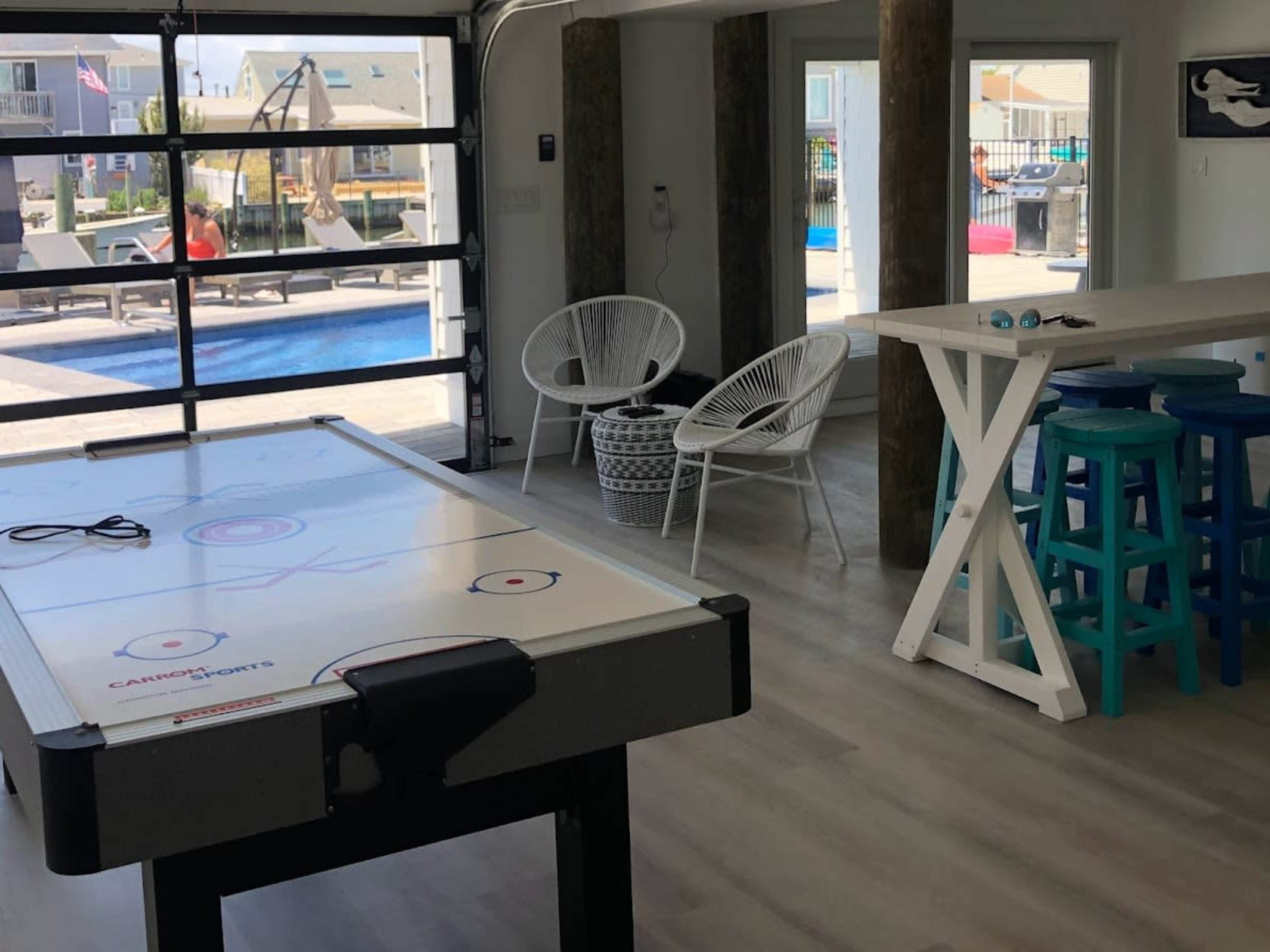 The image shows an indoor space featuring an air hockey table, two white chairs, a long table with colorful stools, and large glass doors leading to an outdoor area with a pool.