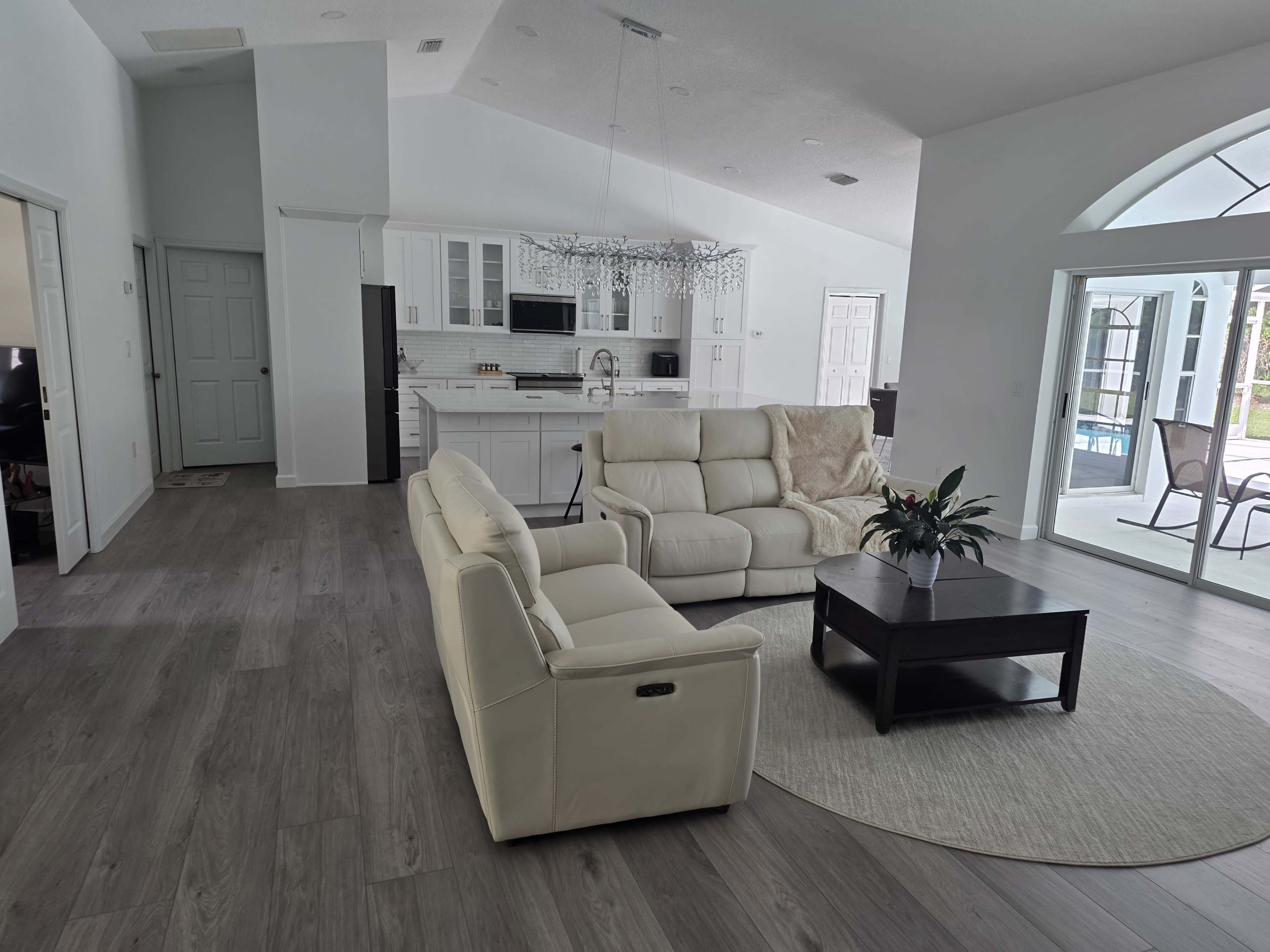 The image shows a modern living room with light-colored furniture, a round area rug, and a view of an adjoining kitchen.