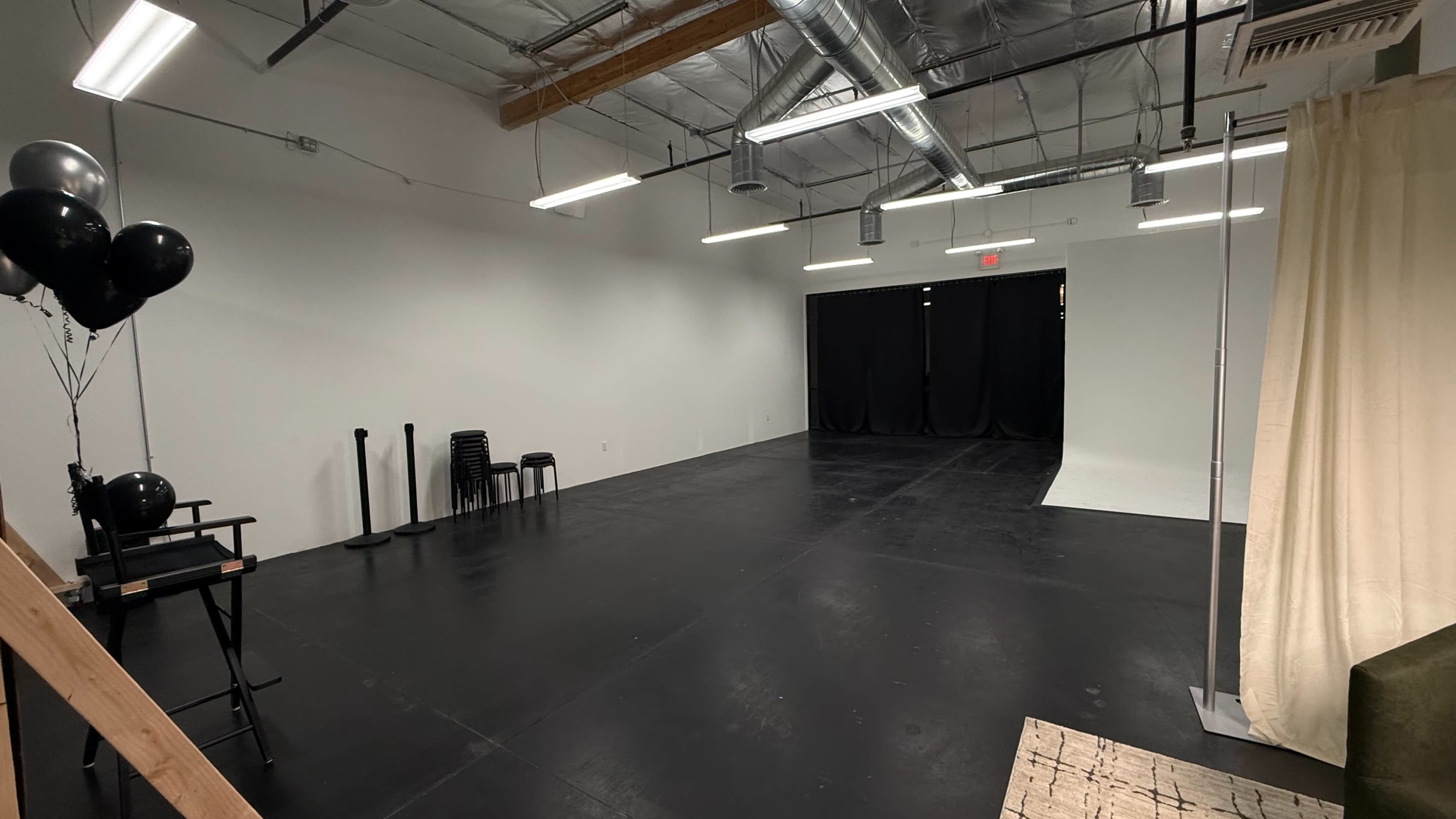 The image shows a spacious empty studio with black flooring, accompanied by a few chairs, a backdrop, and decorative balloons.