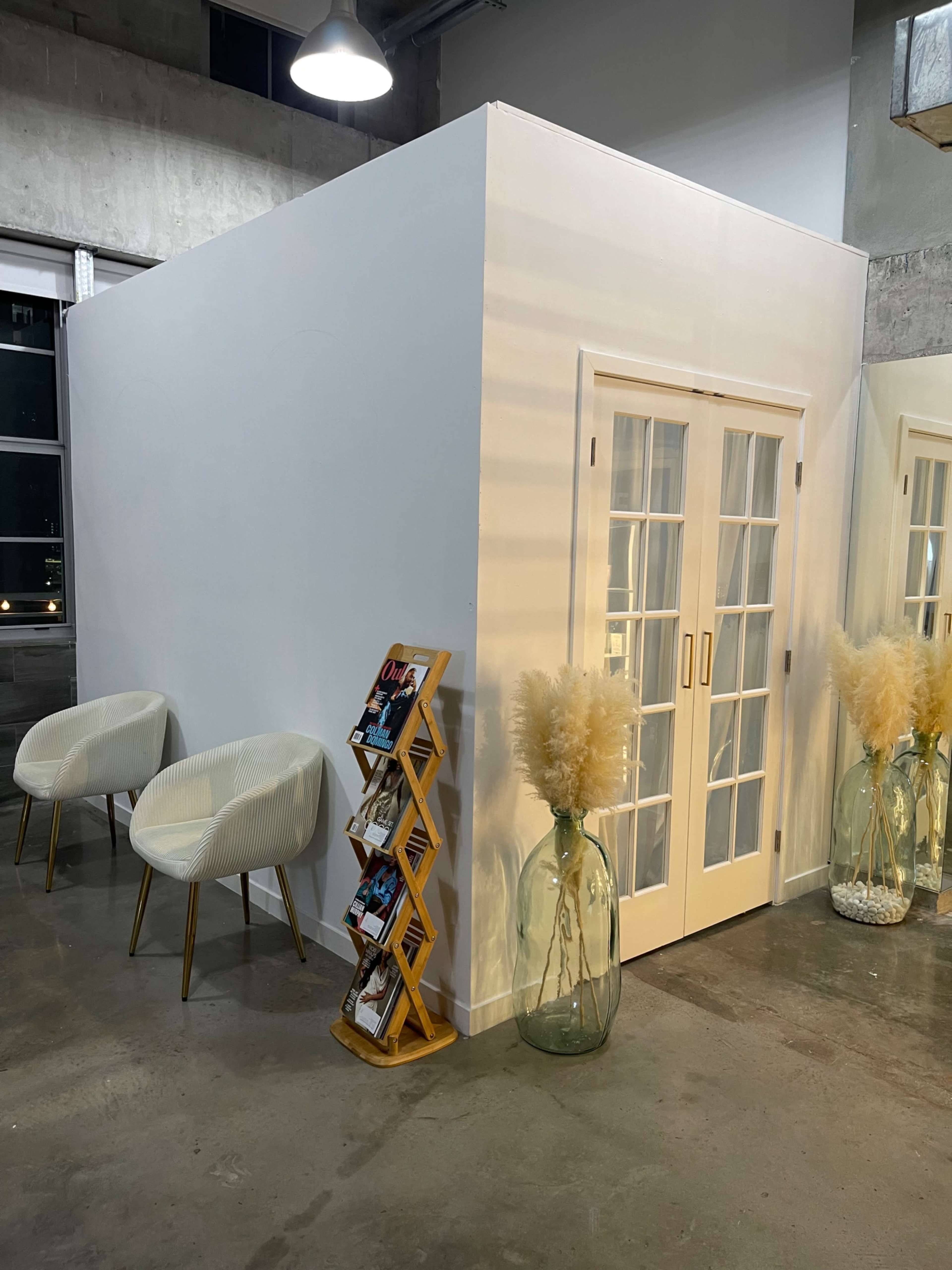 A white-walled room with double doors stands next to a wooden magazine rack and two modern chairs, with large glass vases filled with decorative plants nearby.