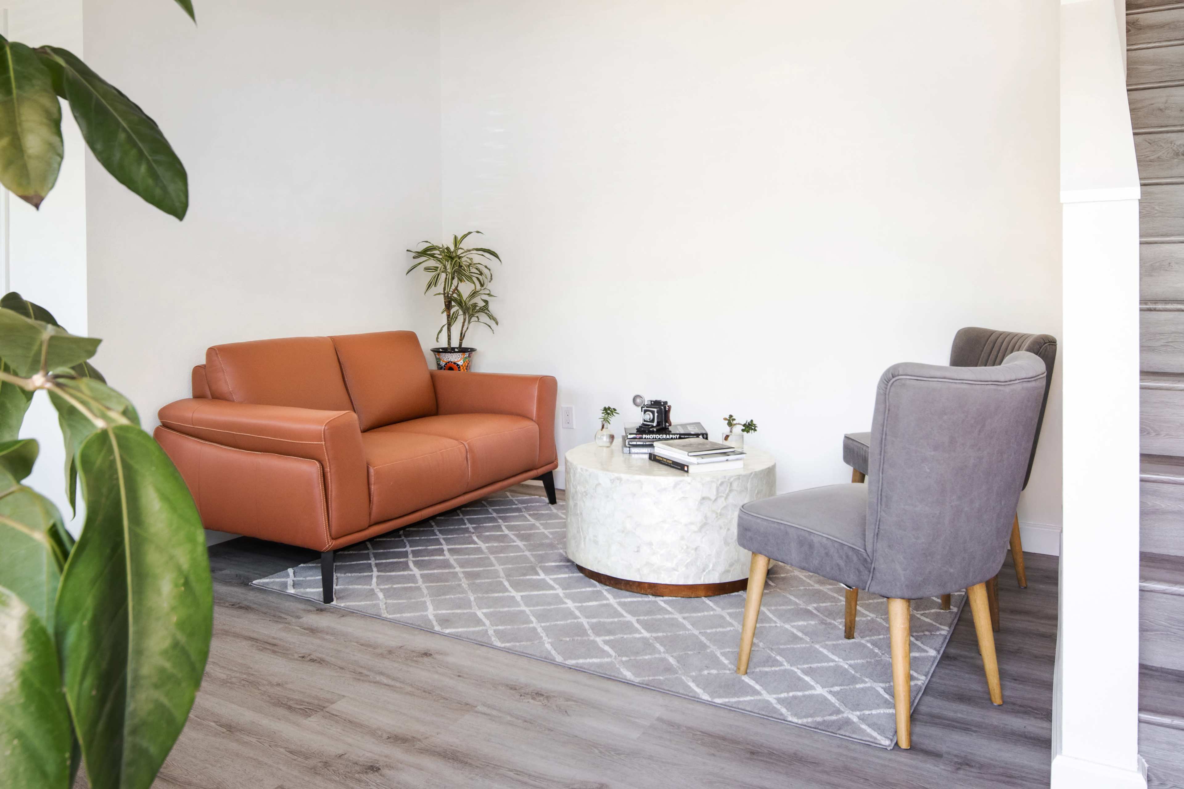 A modern living room features a rust-colored sofa, two upholstered chairs, a round marble coffee table, and a potted plant beside a staircase.