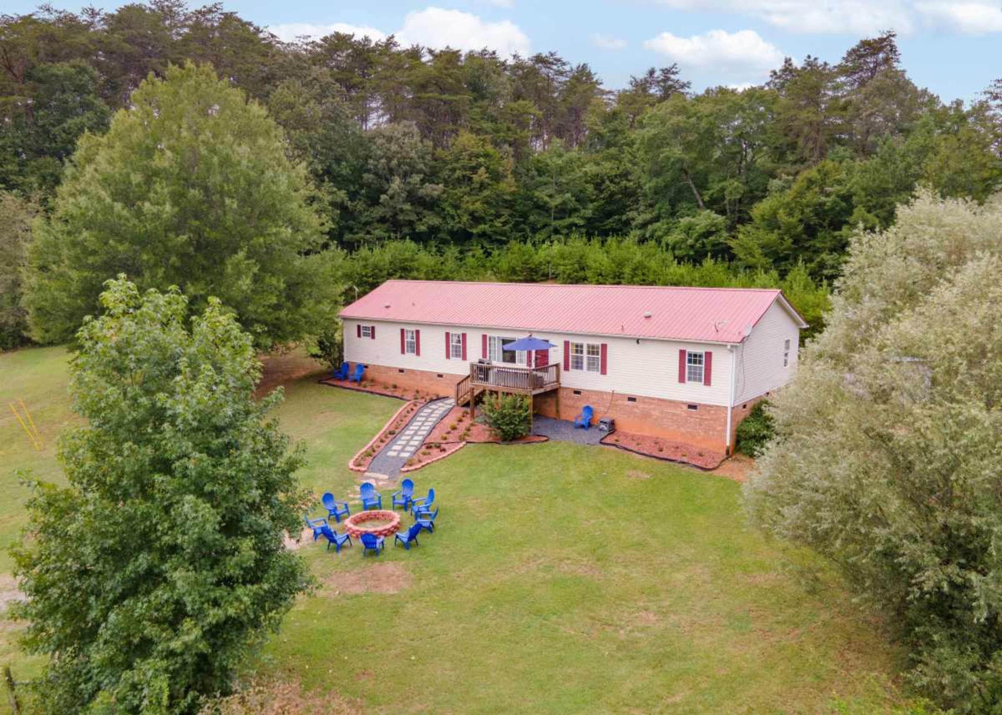 A single-story house with a red metal roof is surrounded by green trees, featuring a landscaped yard with a fire pit and blue seating.
