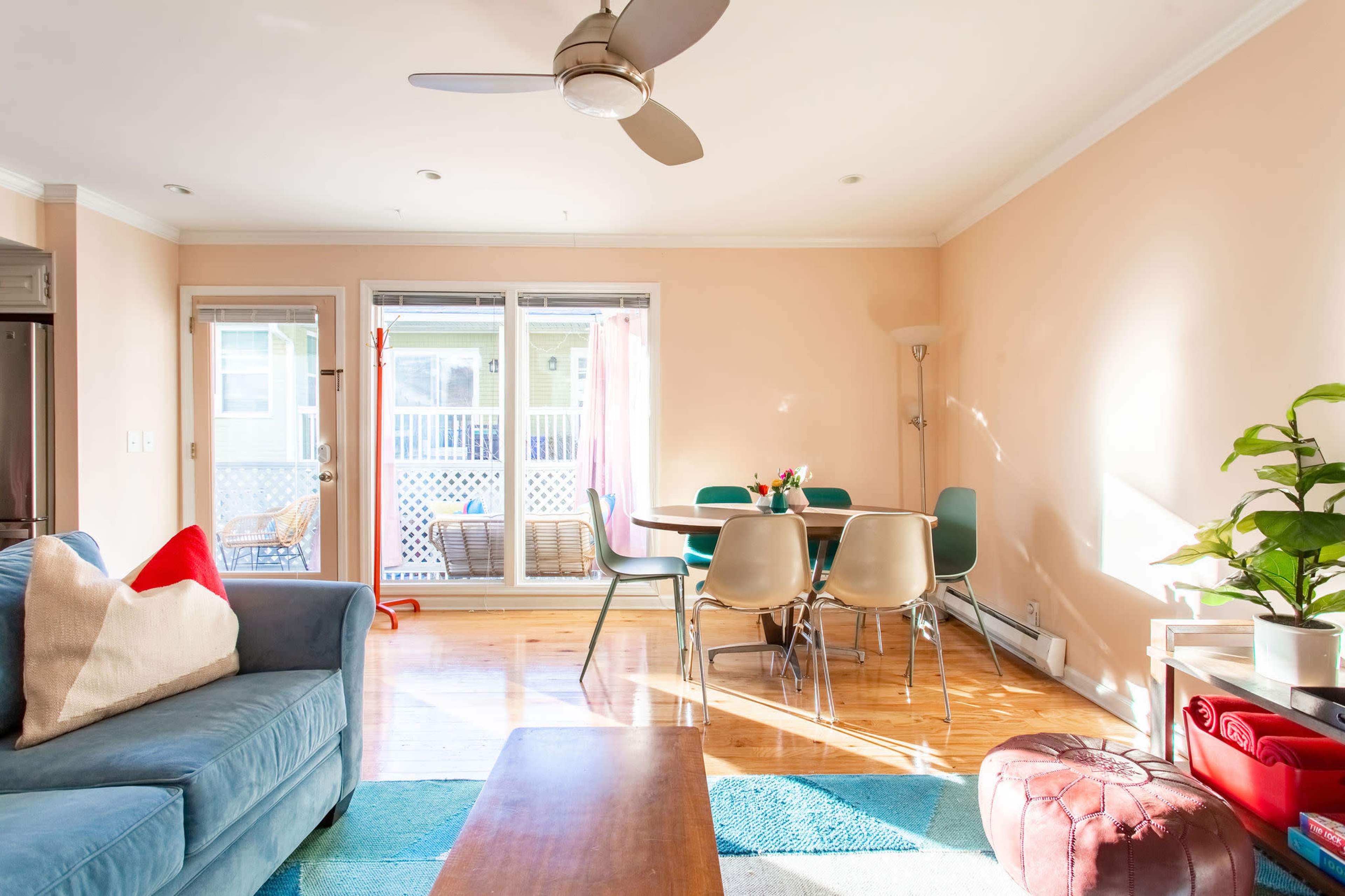 The scene features a living area with a blue couch, a wooden coffee table, a dining table with colorful chairs, and large sliding doors that open to an outdoor patio.