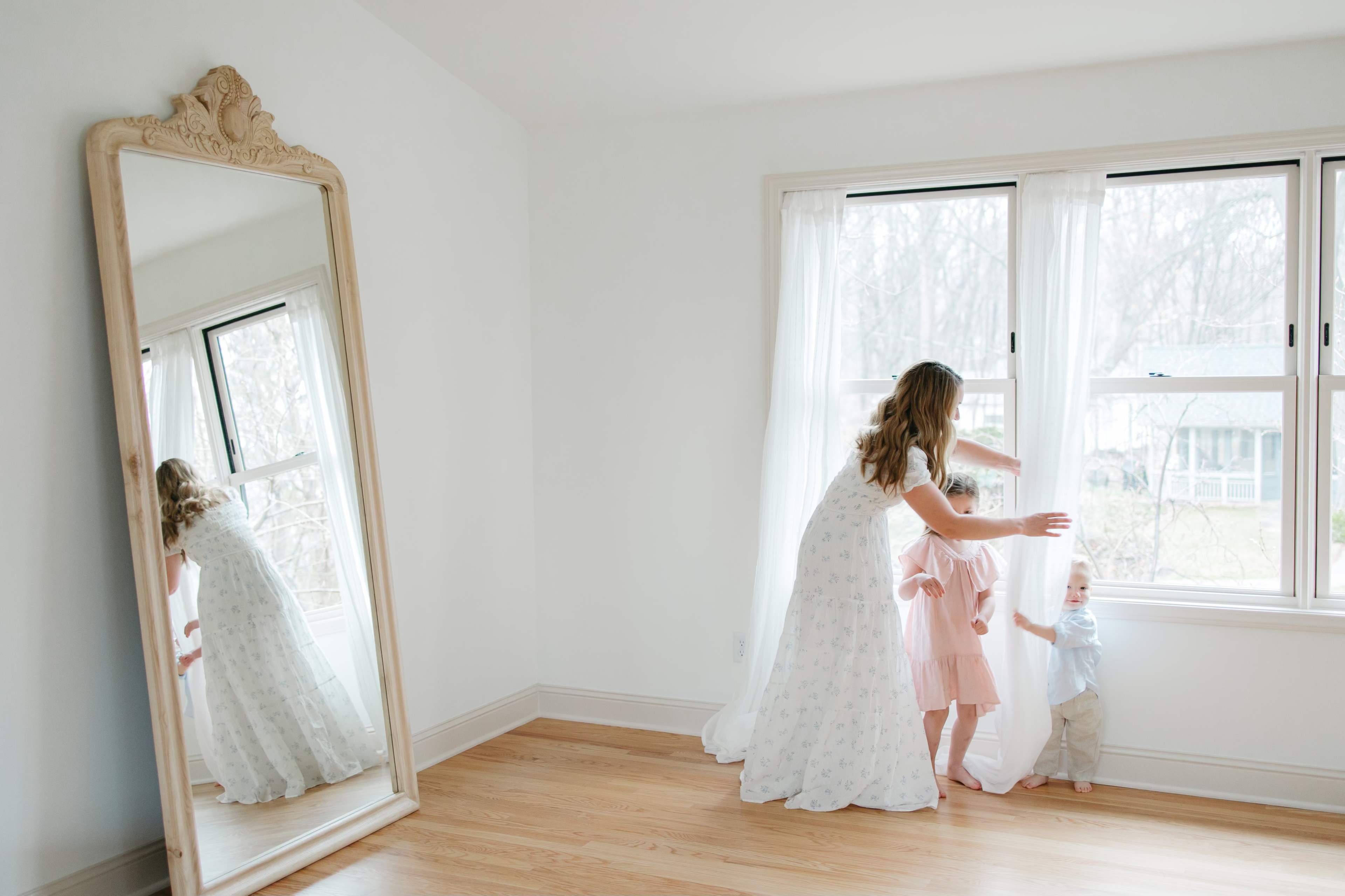 A woman in a white dress assists two children with adjusting sheer curtains beside a large, ornate mirror in a bright room.