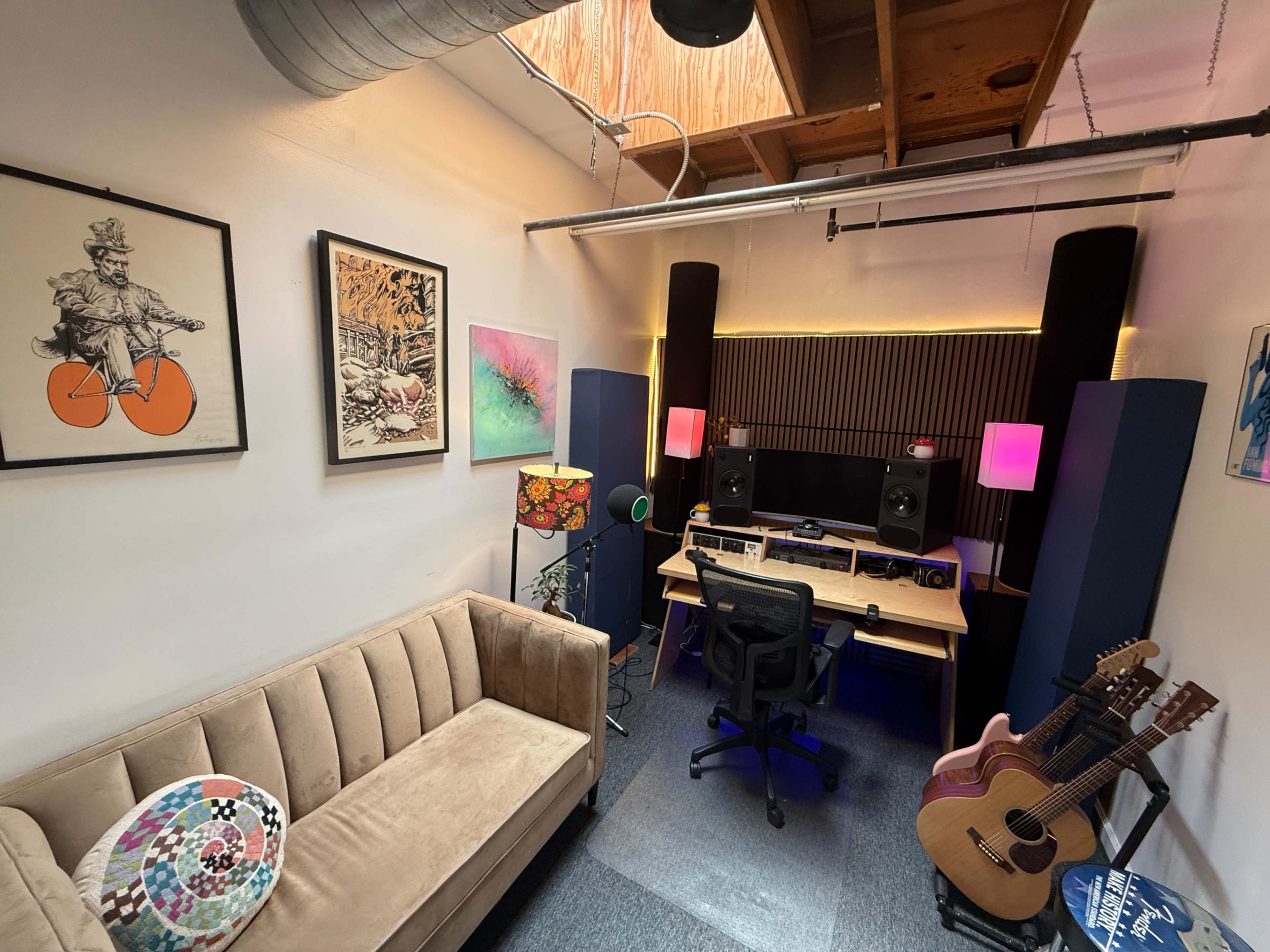 The image shows a cozy music studio with a desk featuring audio equipment, two large speakers, a sofa, and various artworks on the walls.