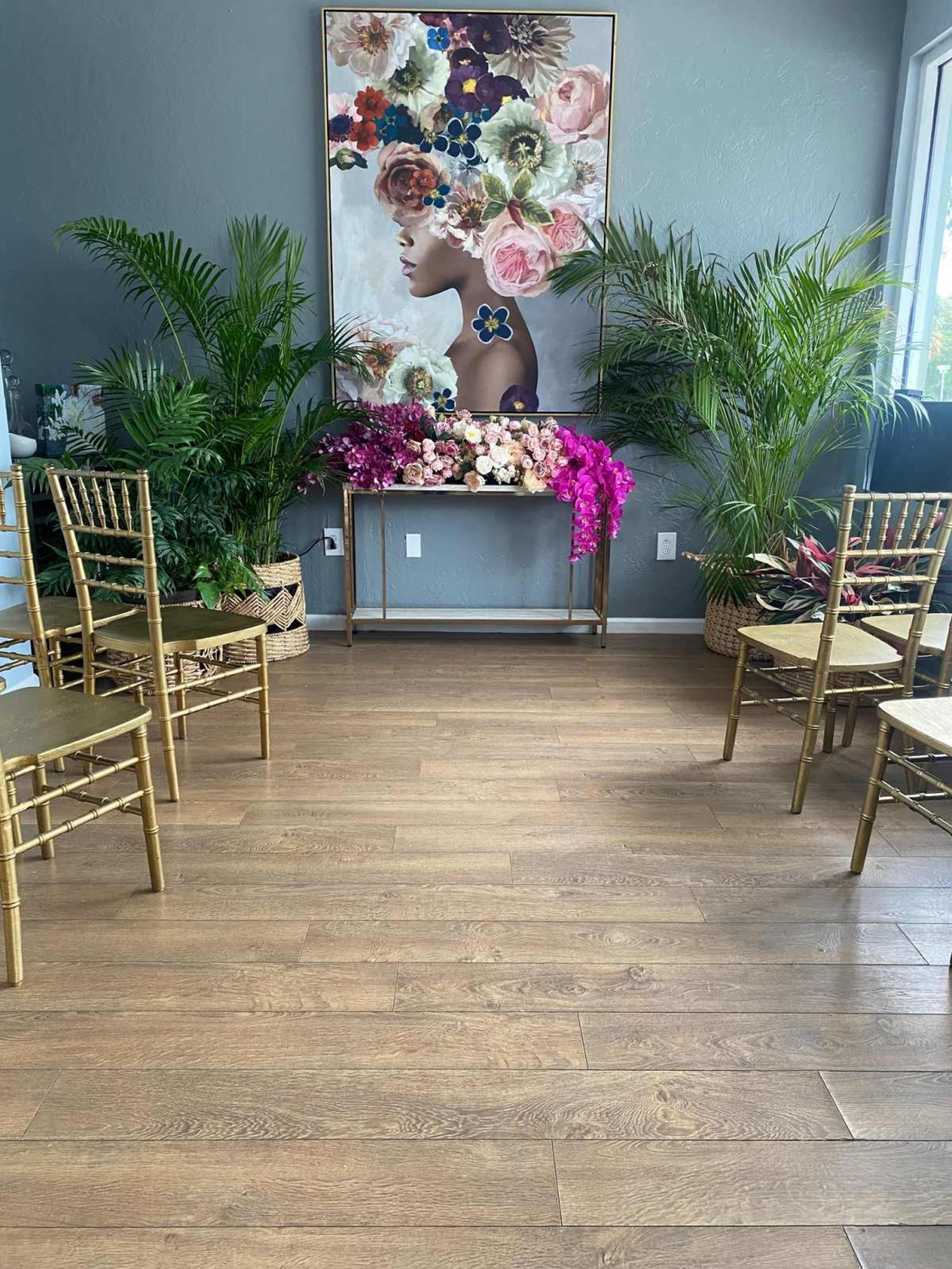 A room features a floral-themed wall art, a floral display on a table, and four golden chairs arranged in front of it, surrounded by potted plants.