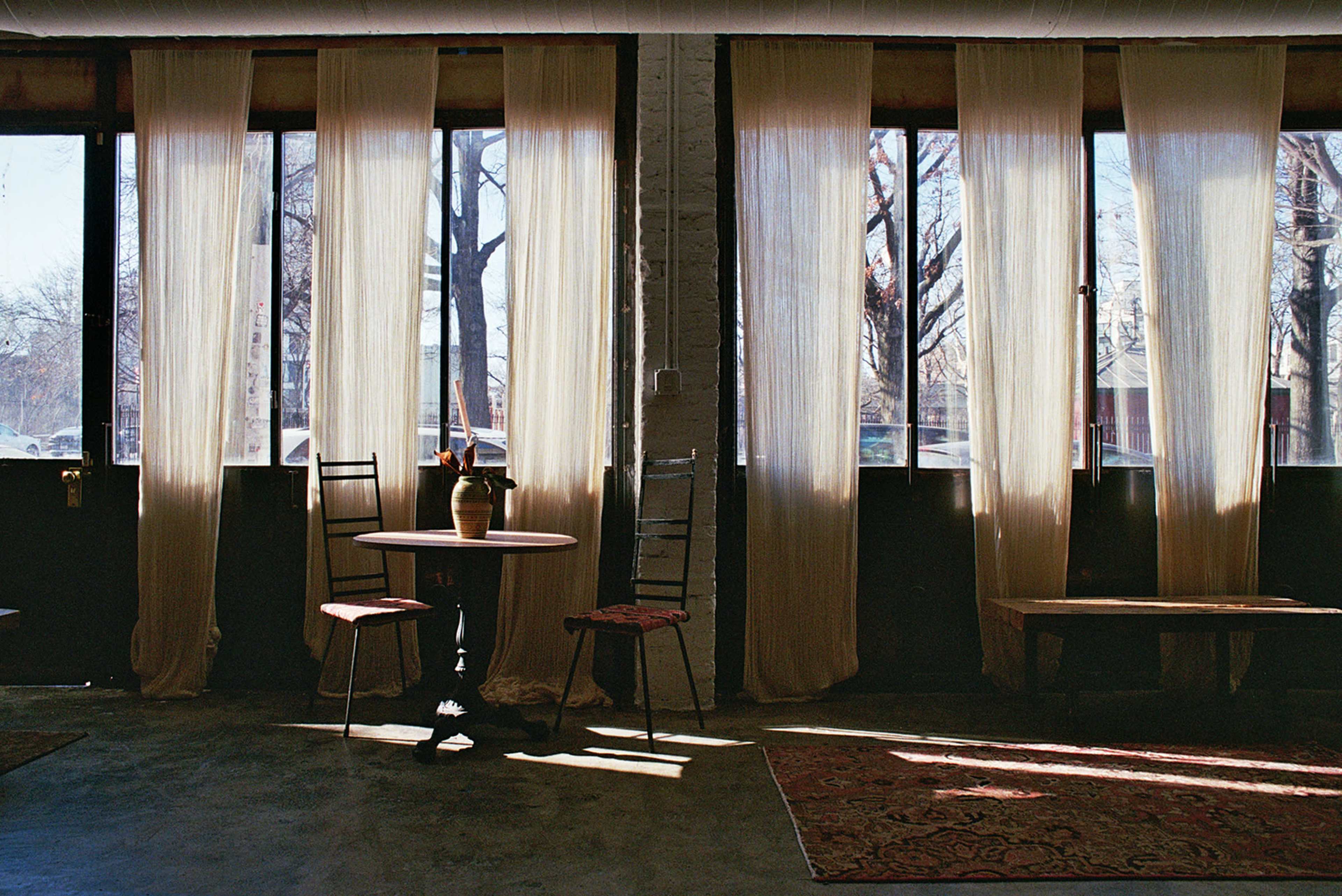 A small round table with a vase sits between two tall windows covered by sheer curtains, illuminating a space with a rustic floor and simple chairs.