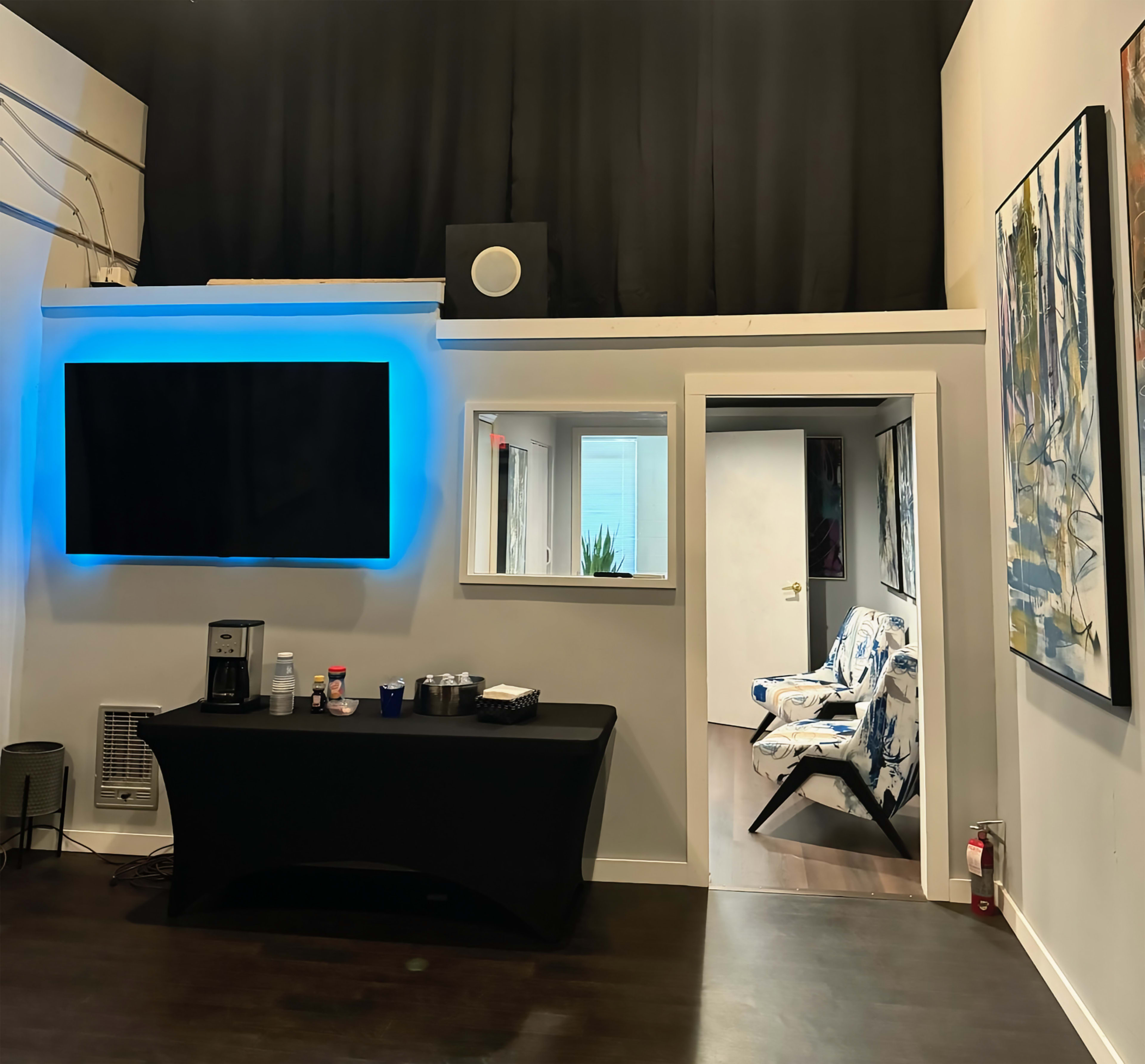 The image shows a modern room with a black table displaying drinks, a large TV on the wall with blue lighting, and a doorway leading to another area featuring a patterned chair and artwork.