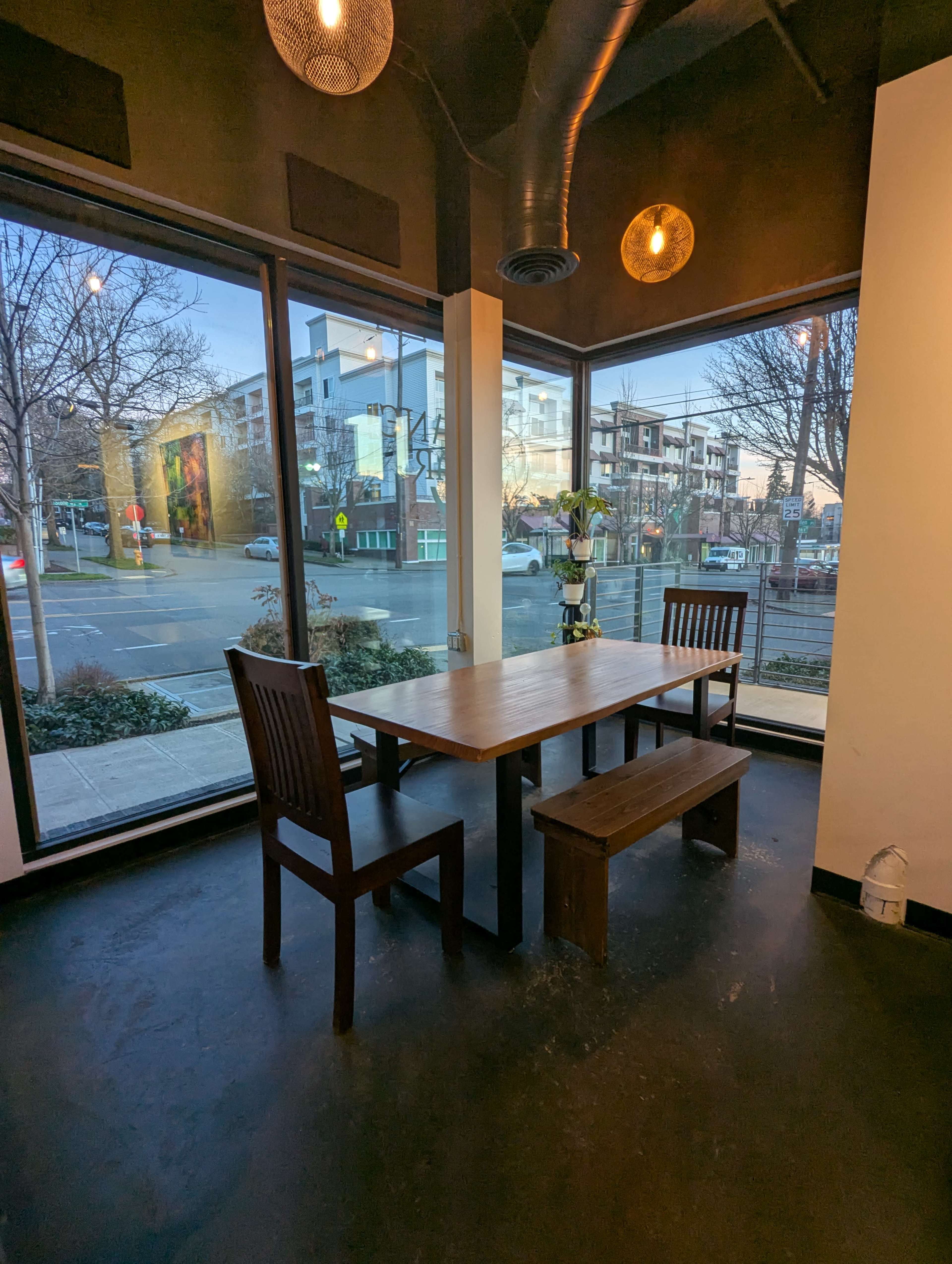 A wooden table with chairs and a bench is positioned near large windows in a modern dining area, revealing an urban street view outside.