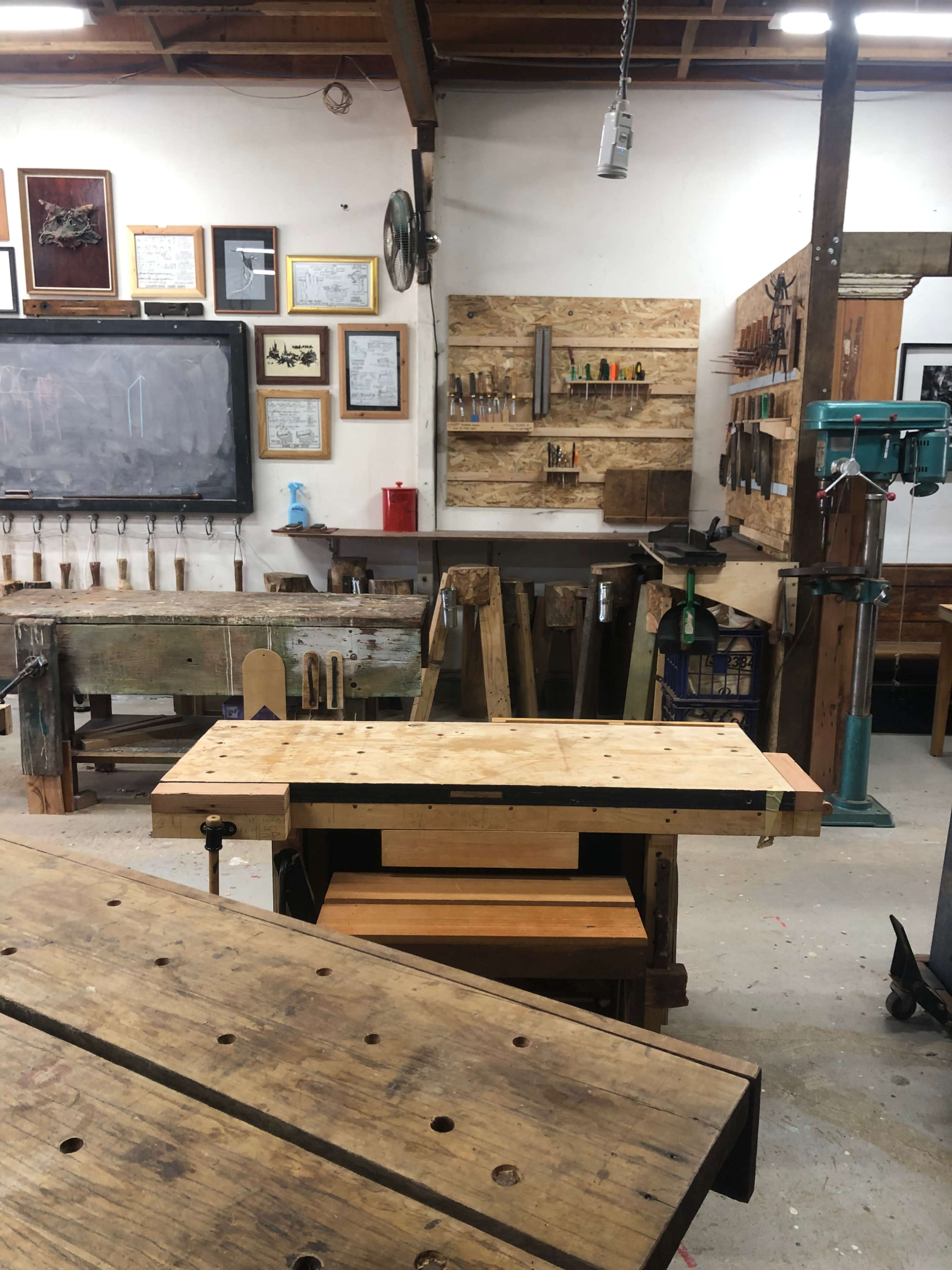 A woodworking shop with wooden workbenches, tools hanging on the walls, and a variety of equipment arranged in a functional layout.