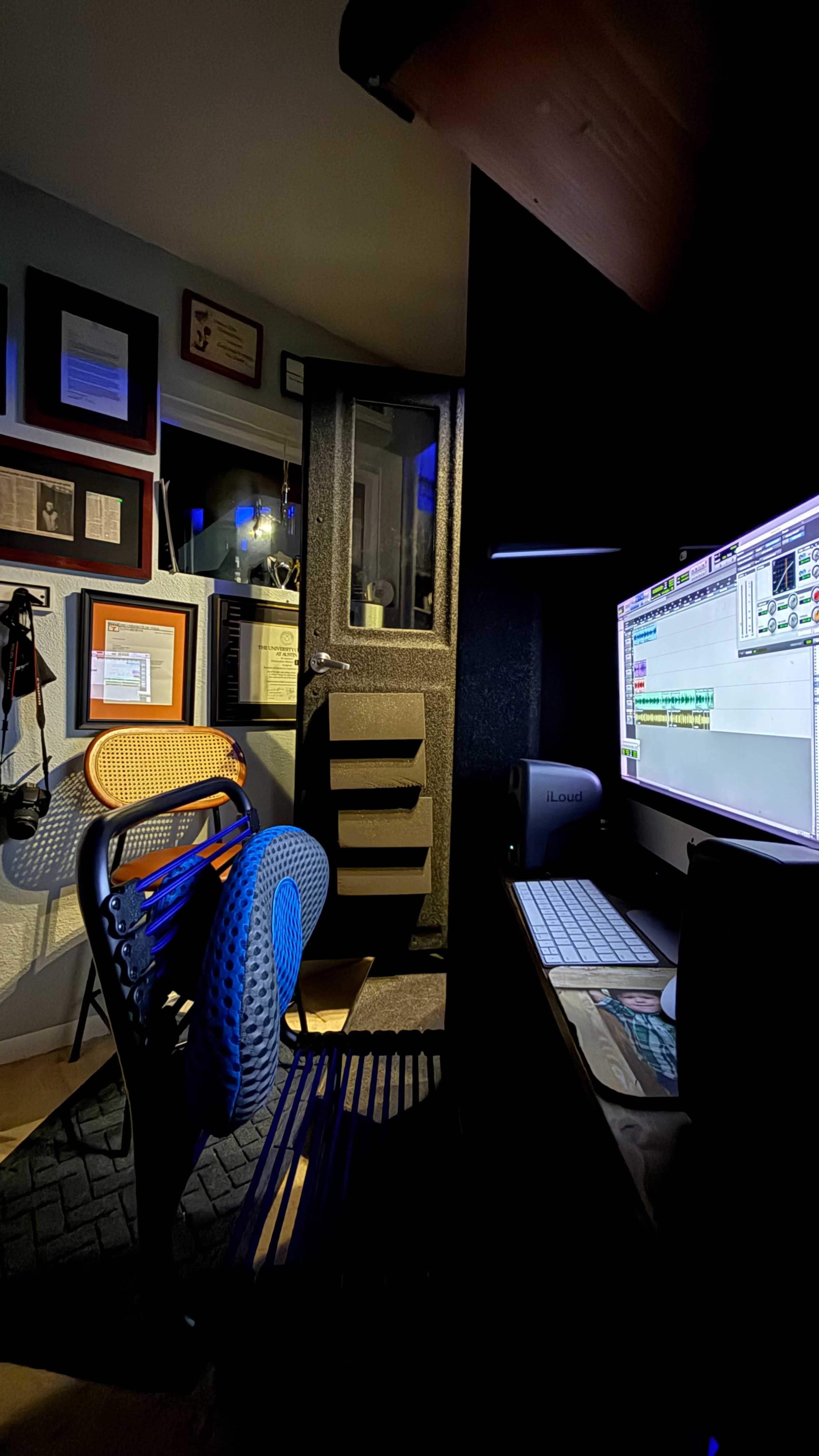 The image shows a dimly lit recording studio featuring a soundproof booth, a computer setup, and various framed certificates on the walls.