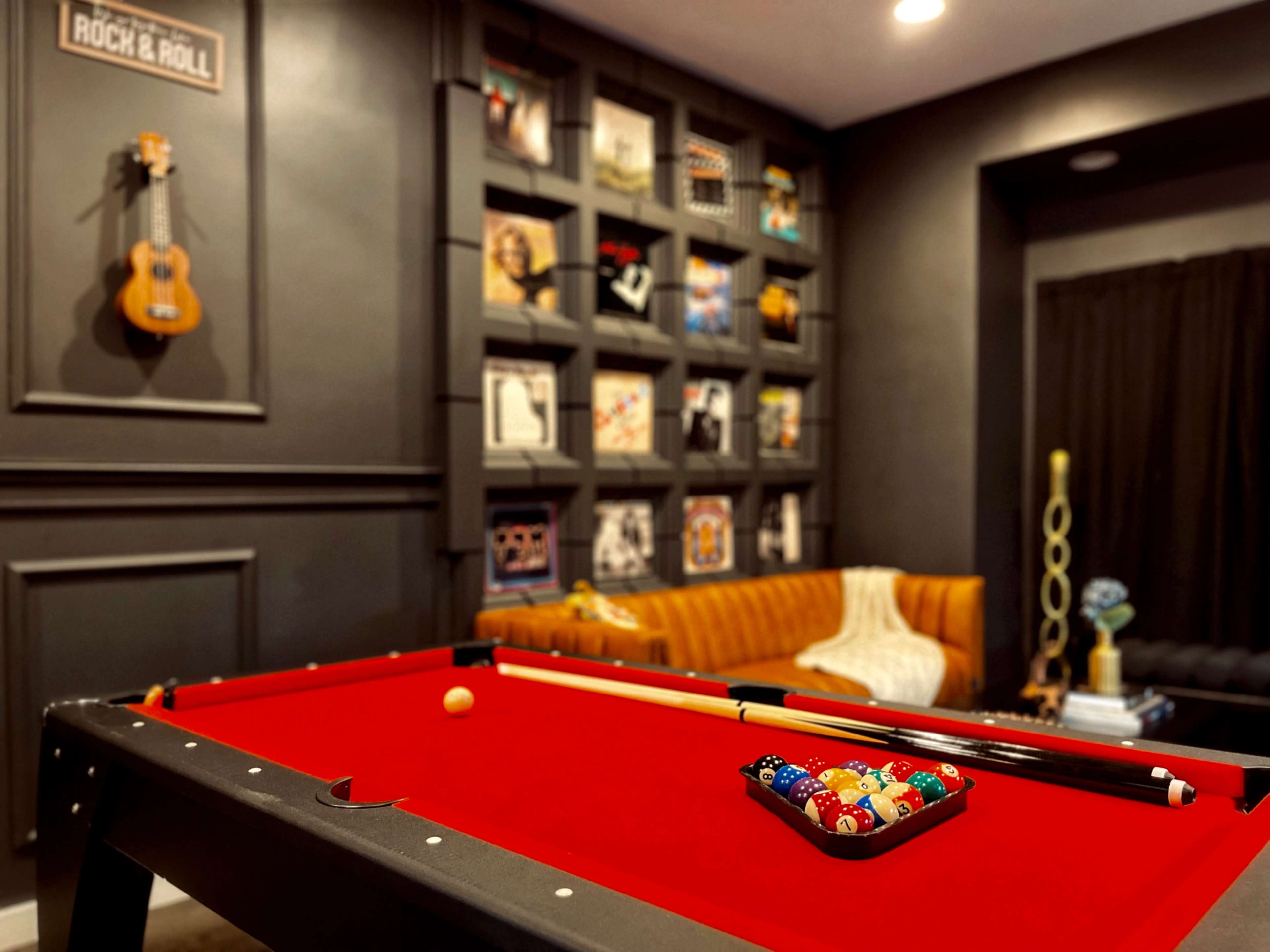 A red pool table with a set of colorful poker chips sits in the foreground of a room adorned with framed artwork and a brown leather couch.