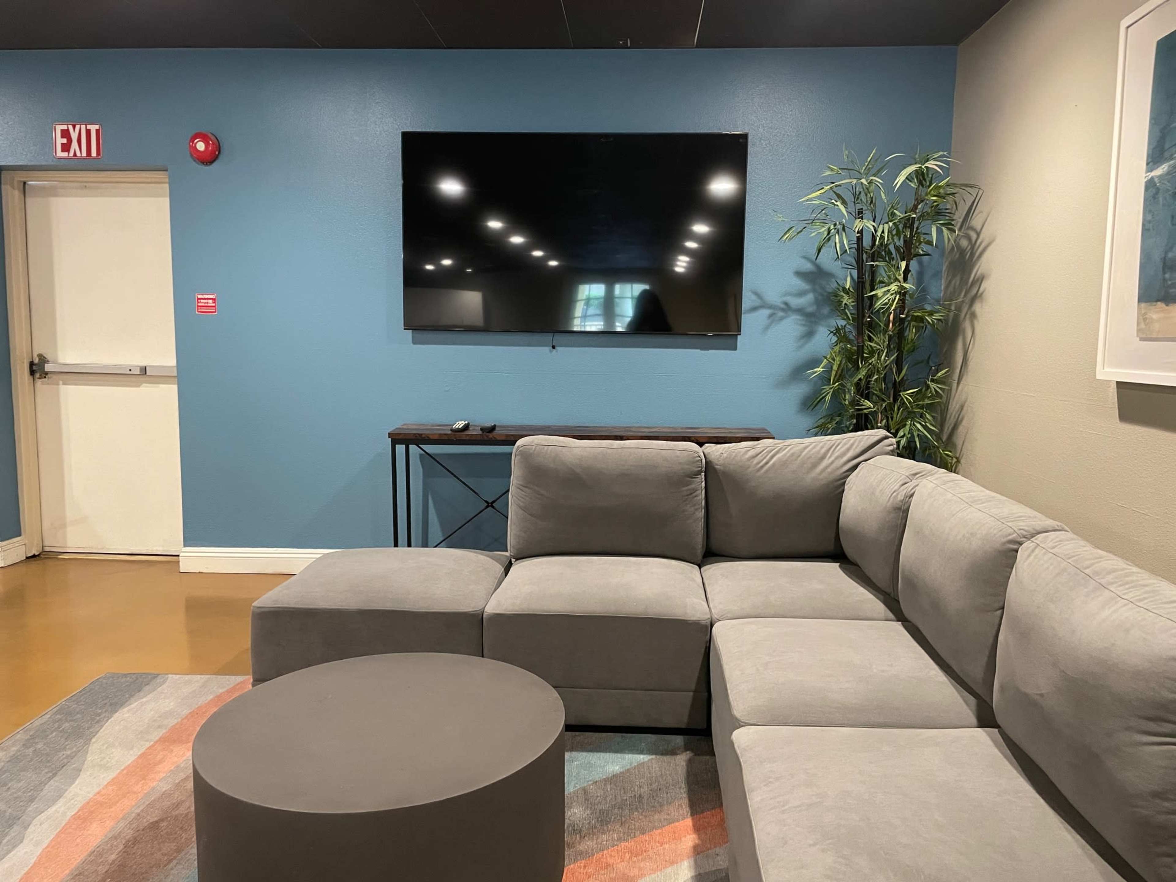A modern lounge area features a gray sectional sofa, a round coffee table, and a wall-mounted television against a blue backdrop.
