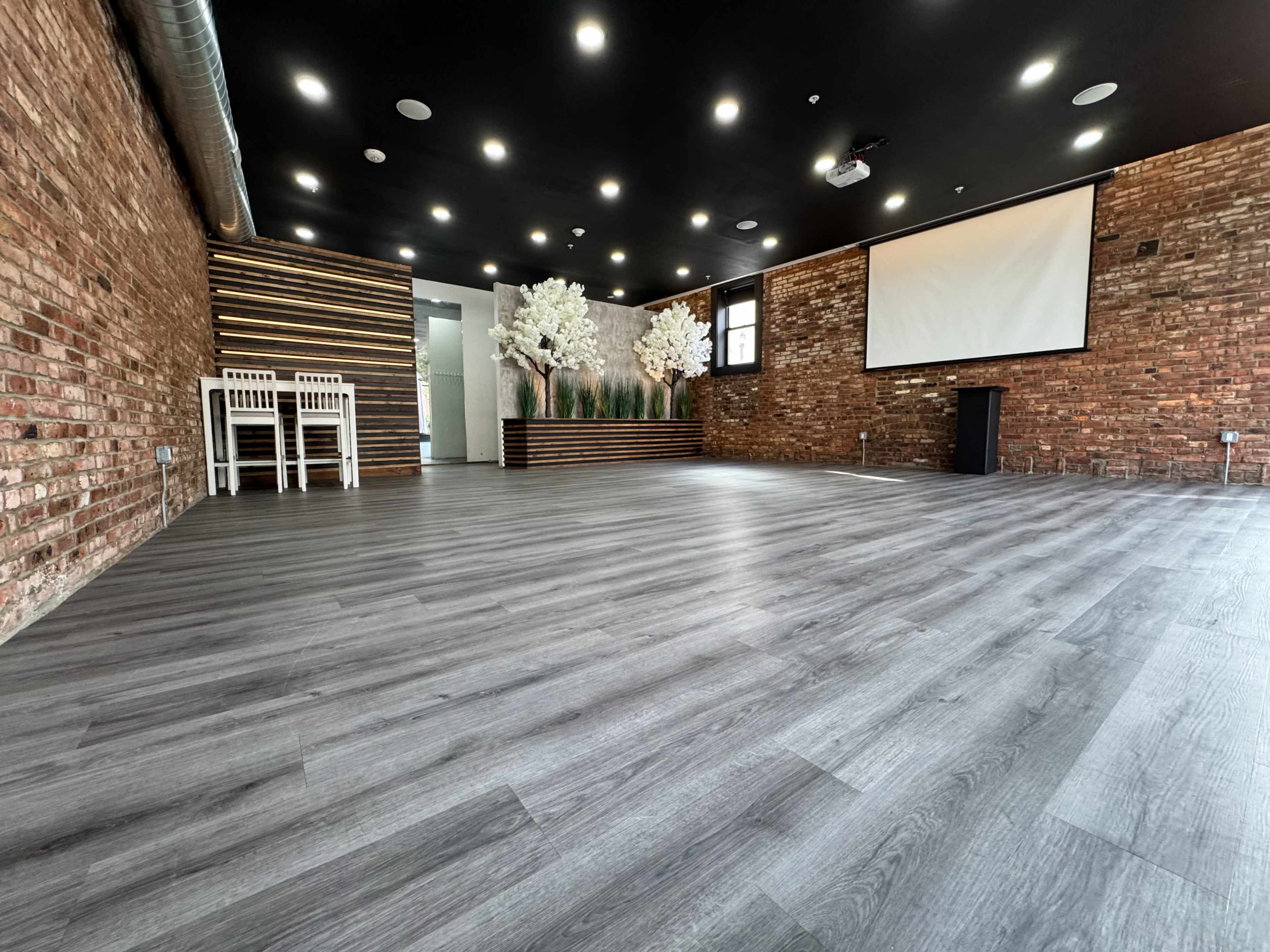 The image shows a spacious interior room with brick walls, a wooden accent wall, a projector screen, and white chairs near the entrance.