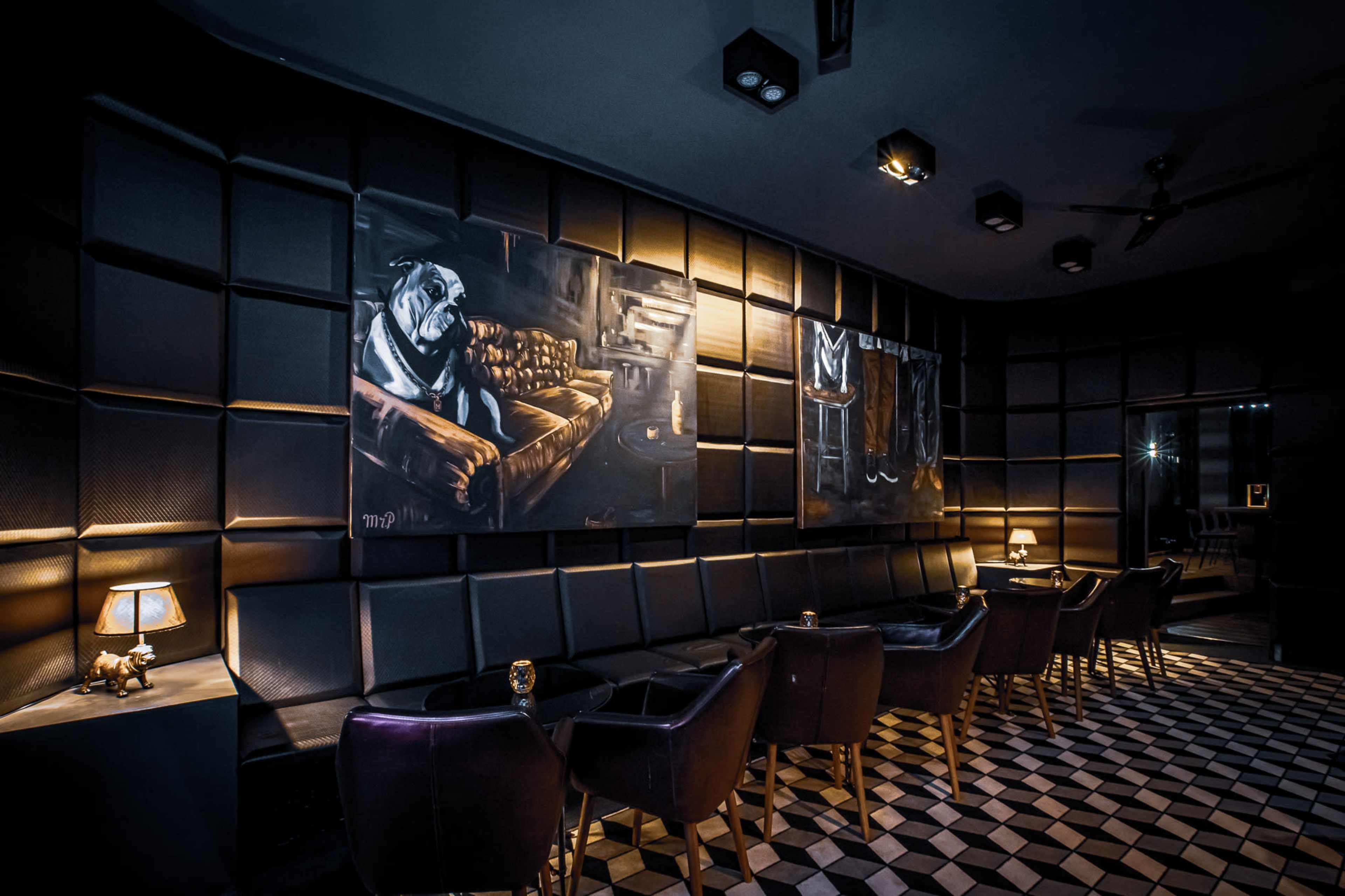 The image shows a dimly lit lounge with black leather seating, a patterned floor, and two large paintings on the wall, one featuring a bulldog.