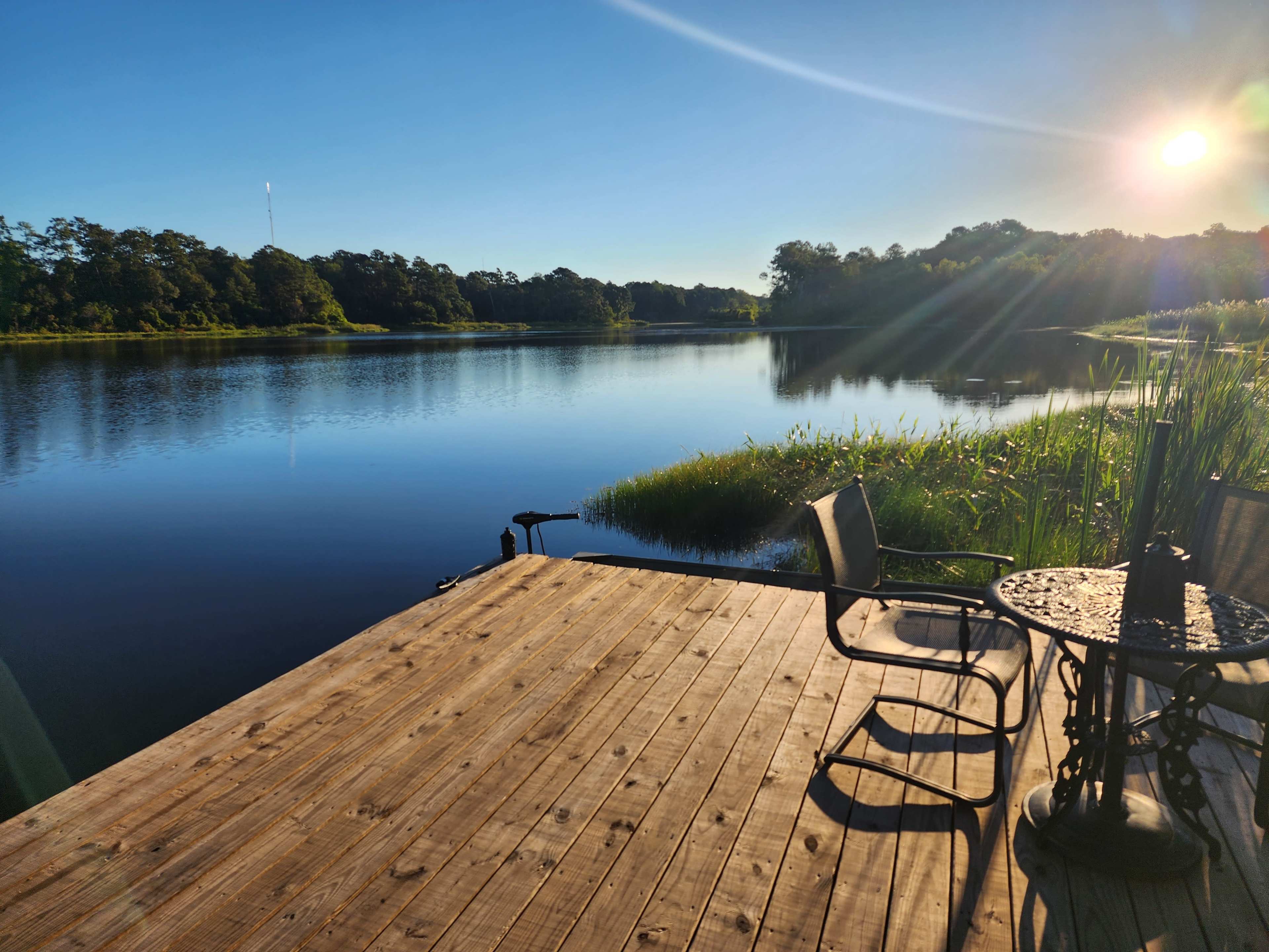 A wooden dock extends over a calm lake surrounded by greenery, with a table and chairs positioned nearby under the bright sun.