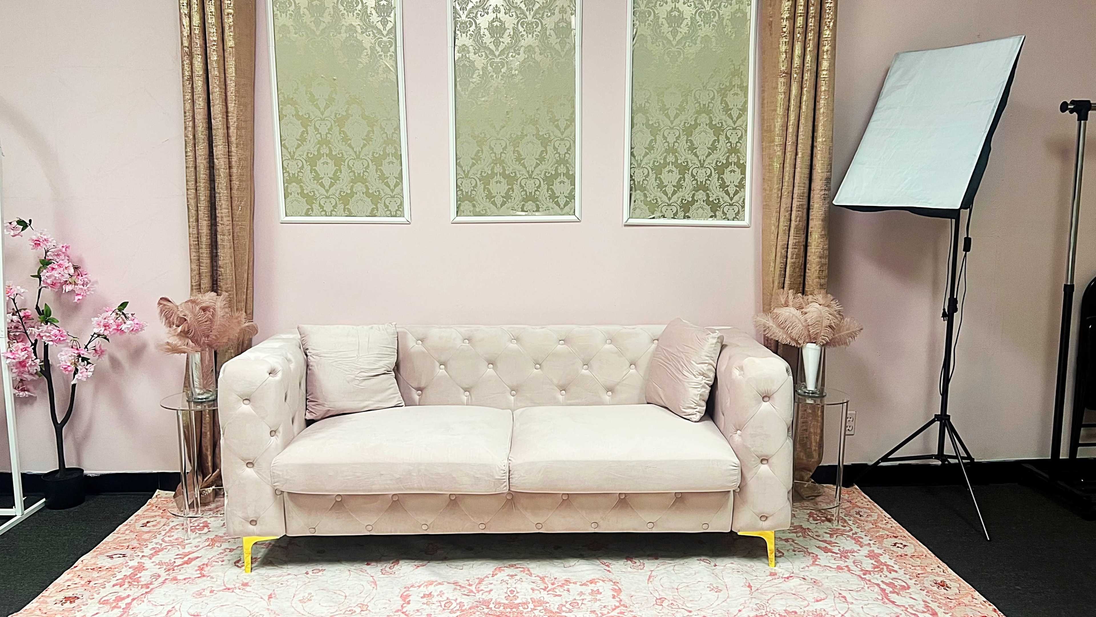The image shows a light pink tufted sofa placed against a pink wall with decorative panels, accompanied by floral arrangements and a photography light setup nearby.
