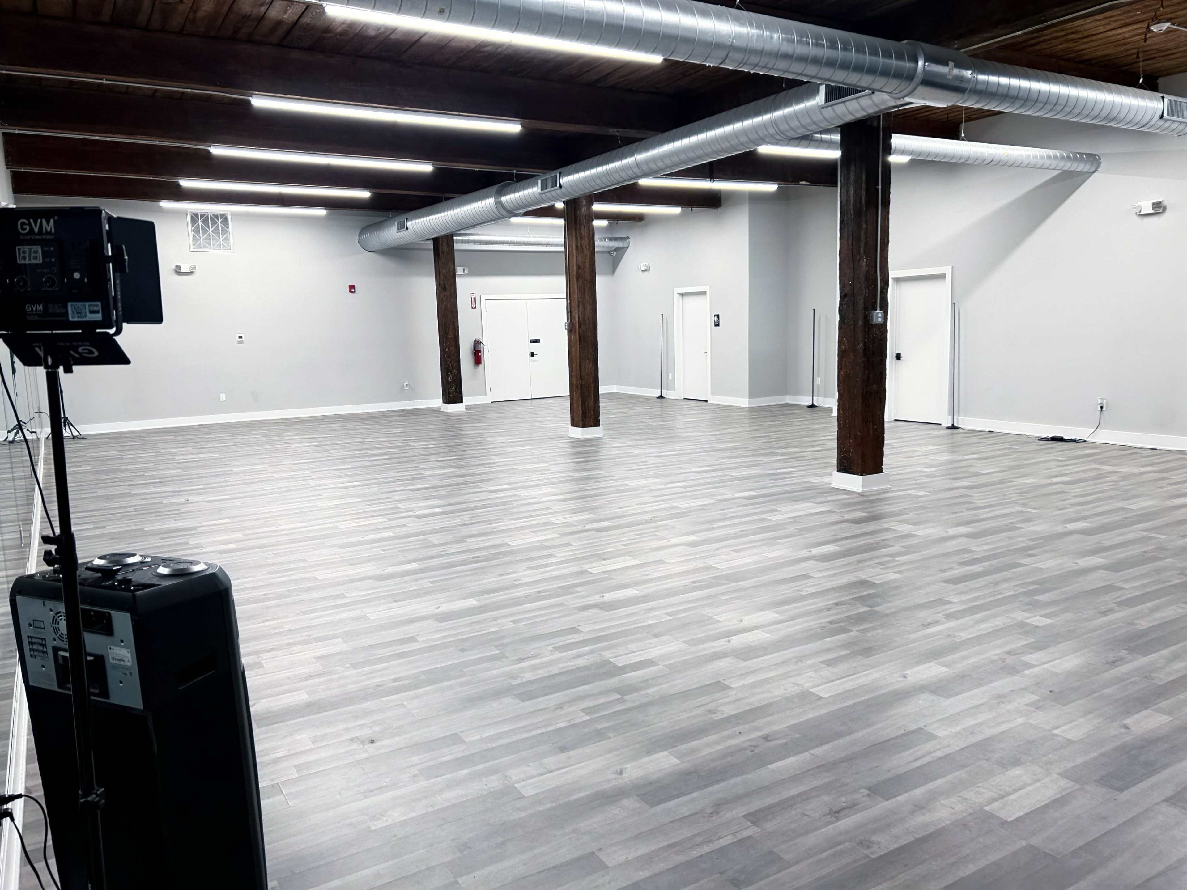 The image shows a large, empty room with gray flooring, white walls, and exposed wooden beams, featuring several doors and an air vent.