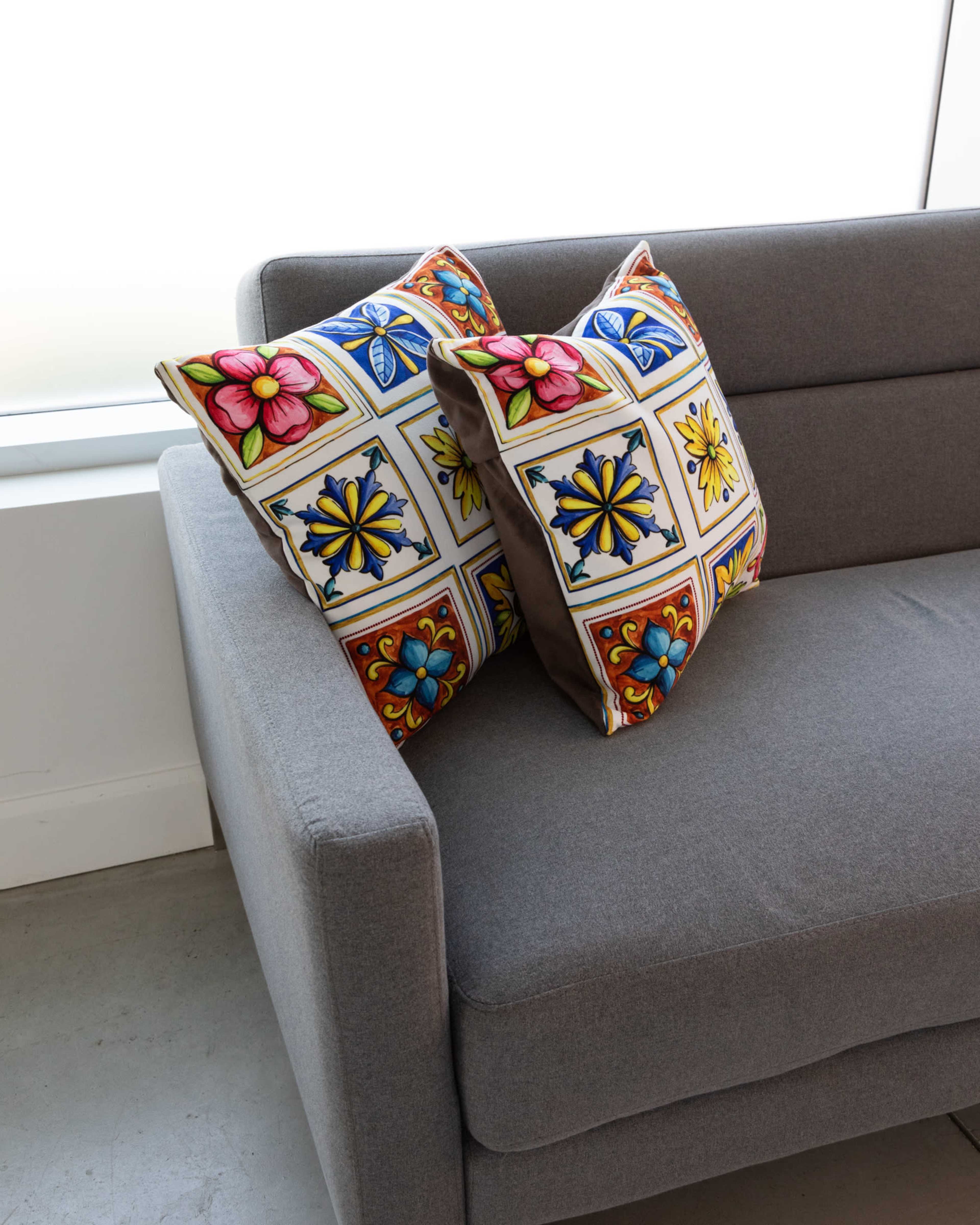 A gray couch with two colorful, patterned pillows is placed near a large window.