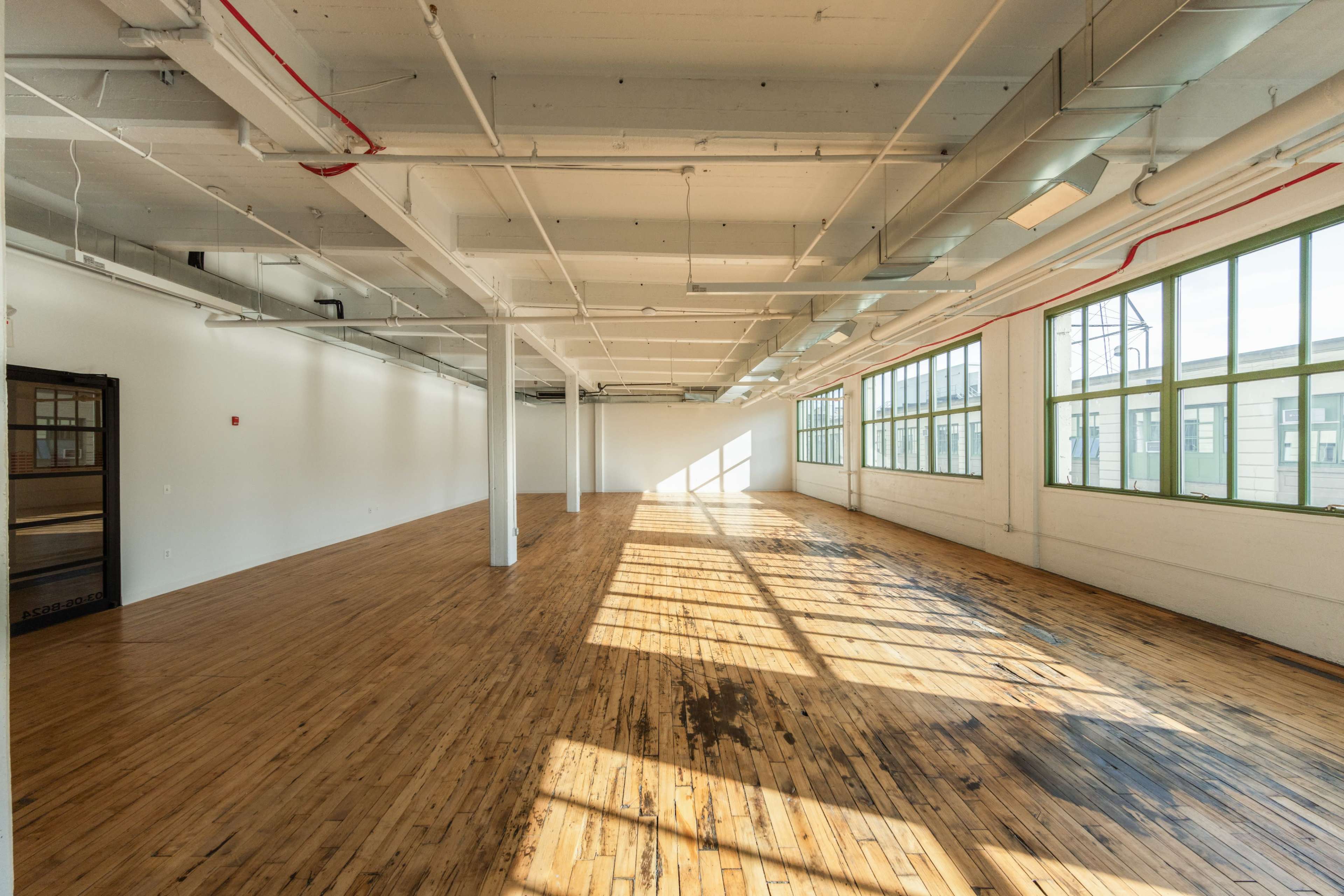 The image shows a spacious, empty room with hardwood floors, large windows, and beams of sunlight illuminating the interior.