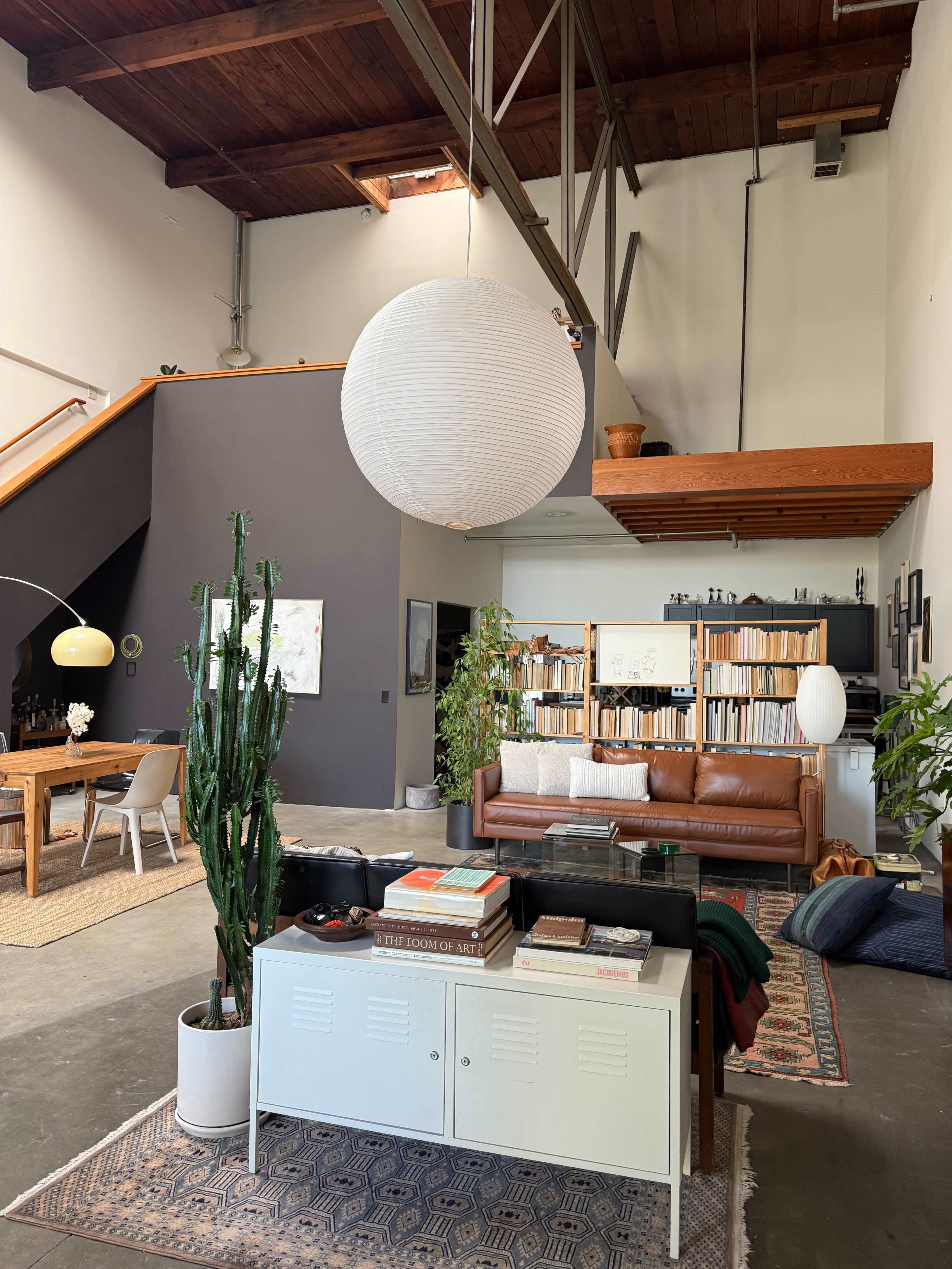 The image features a spacious modern living room with a brown leather sofa, large bookshelves, a white pendant light, and various plants, all set against a backdrop of high wooden ceilings.