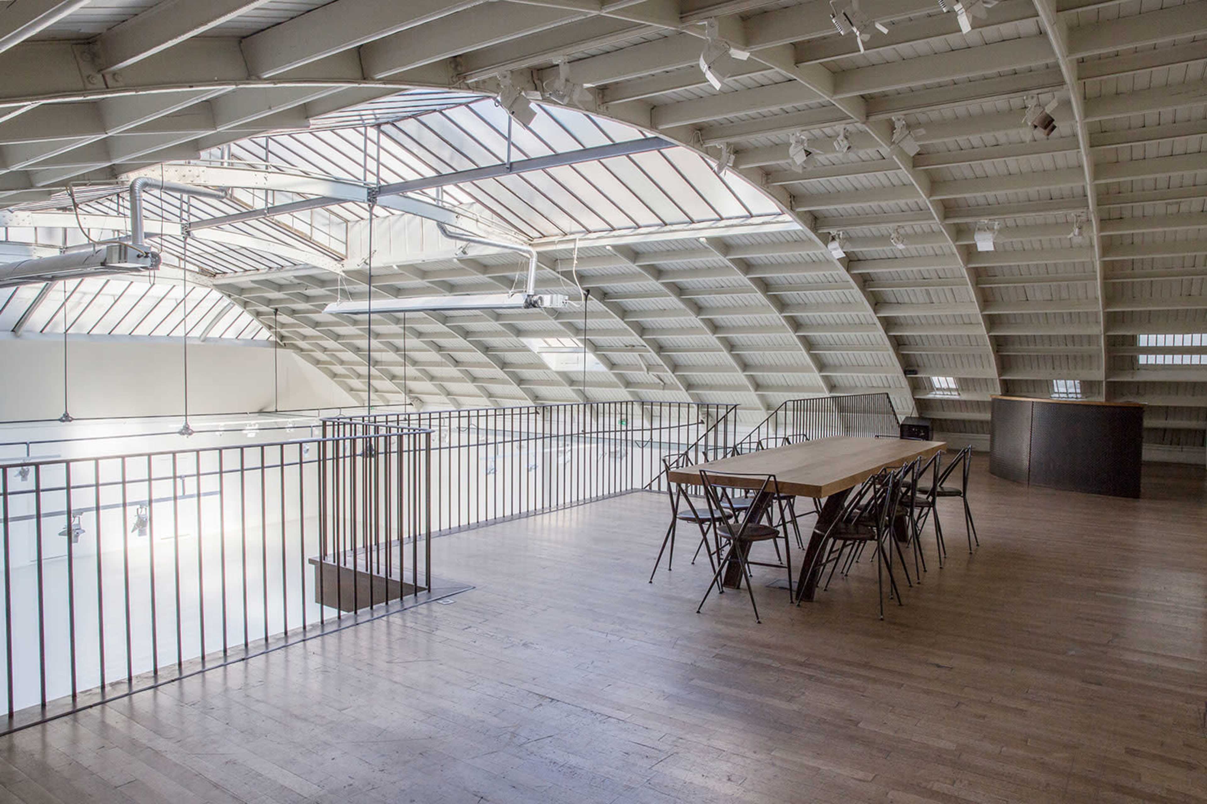 A modern, open loft space with a long wooden table and chairs, featuring a curved roof and large skylights.