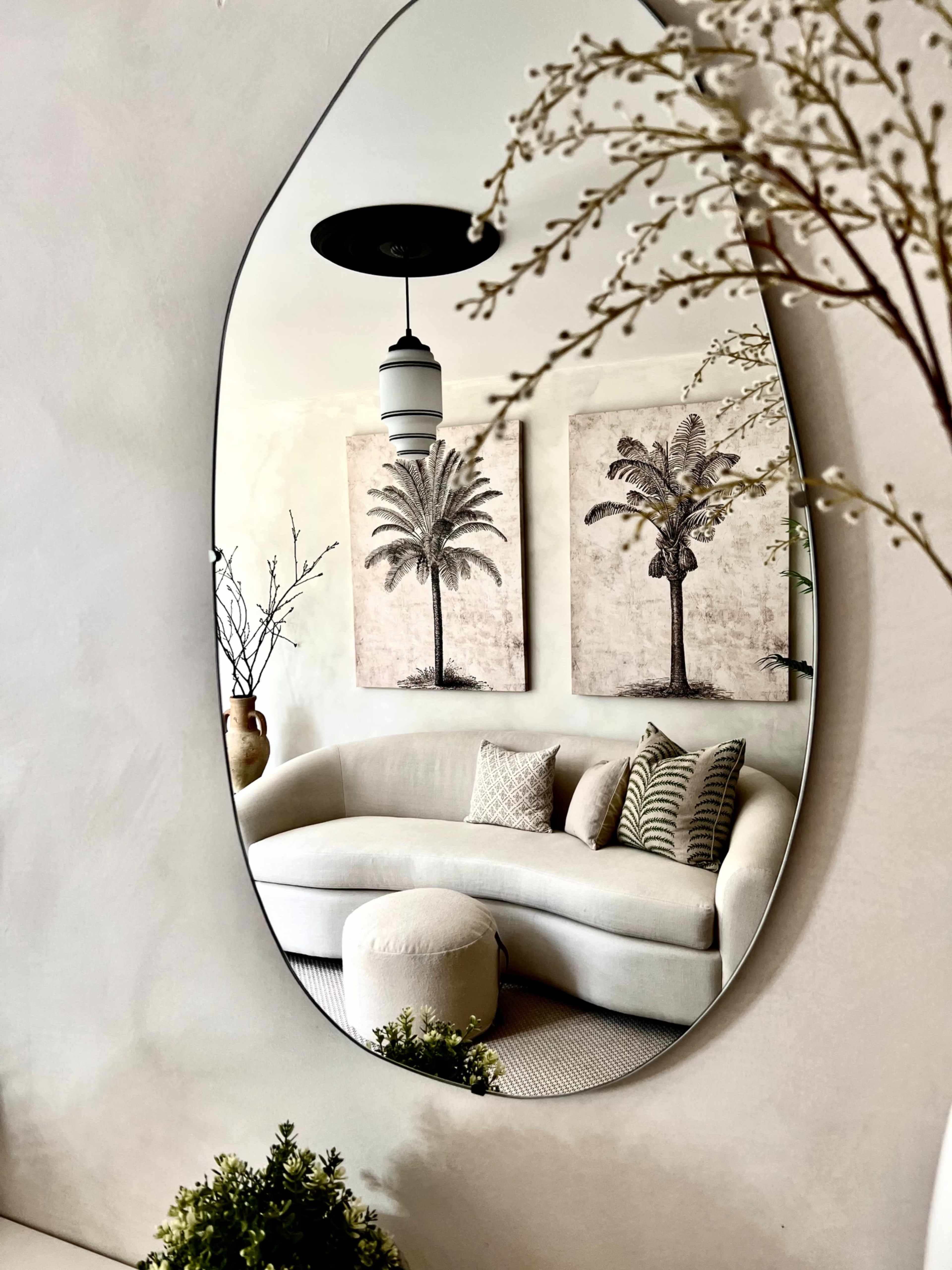 A large oval mirror reflects a cozy living room featuring a curved sofa, decorative pillows, and two palm tree paintings on the wall.