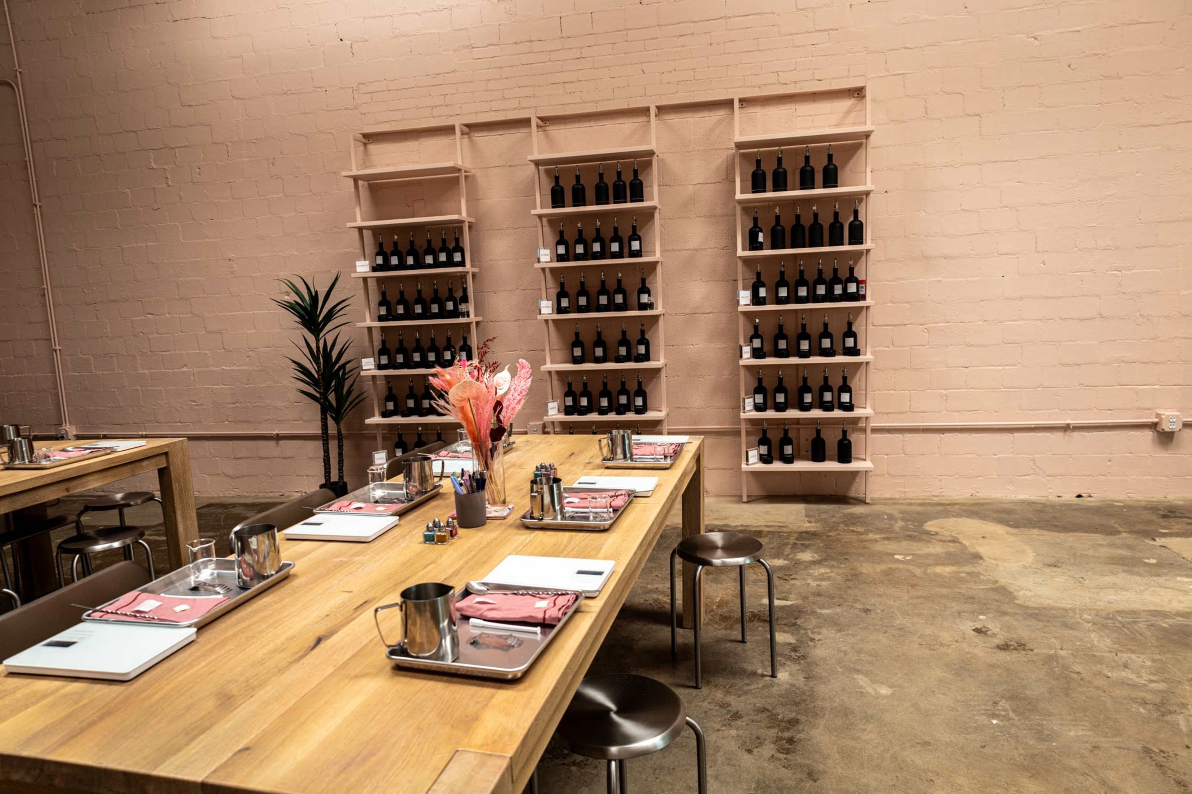 A room with wooden tables and metal stools arranged for a workshop, featuring a wall shelf filled with bottles and decorative elements like a plant and flower arrangement.