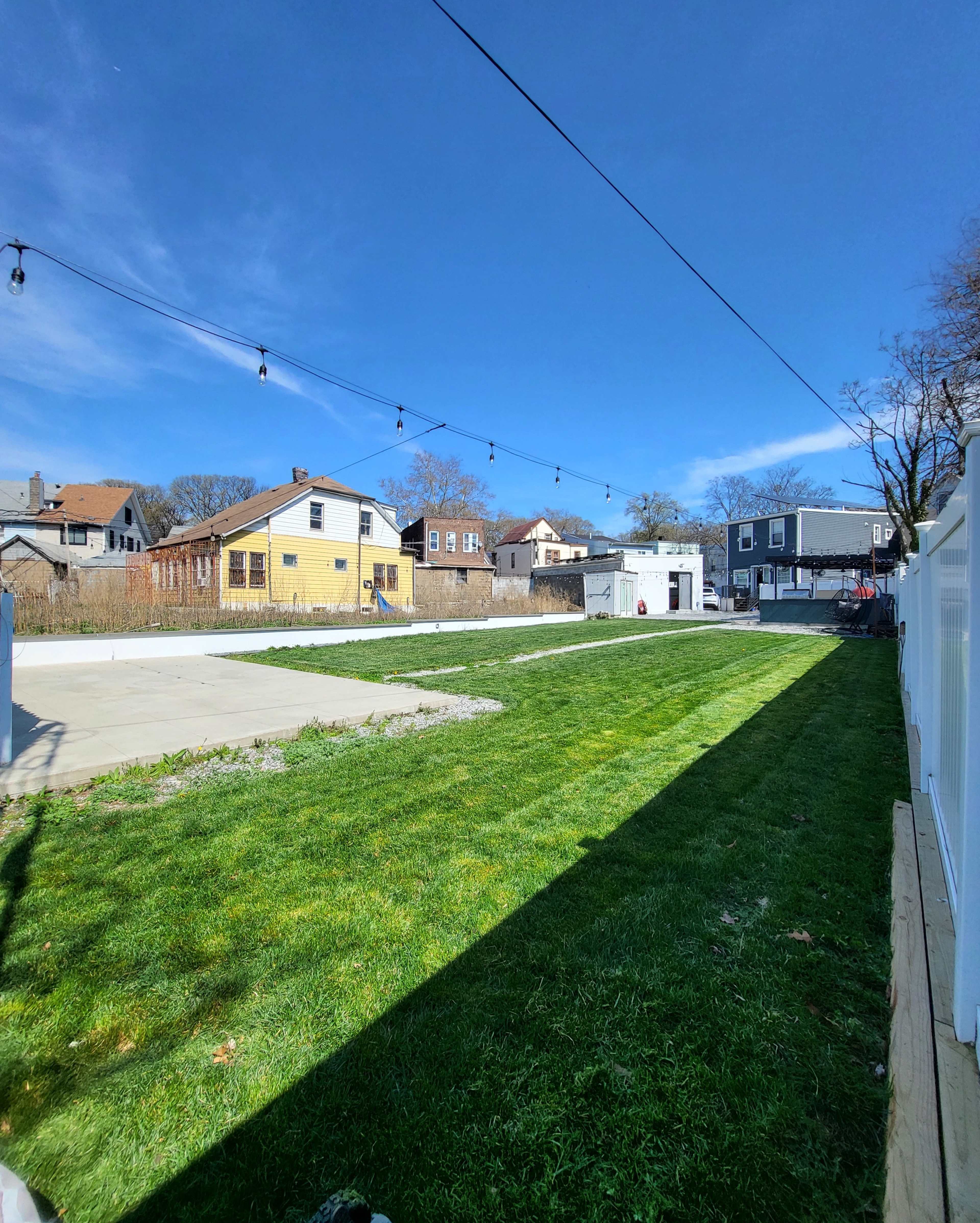 The image shows a grassy backyard area with a concrete surface on one side and several houses in the background under a clear blue sky.