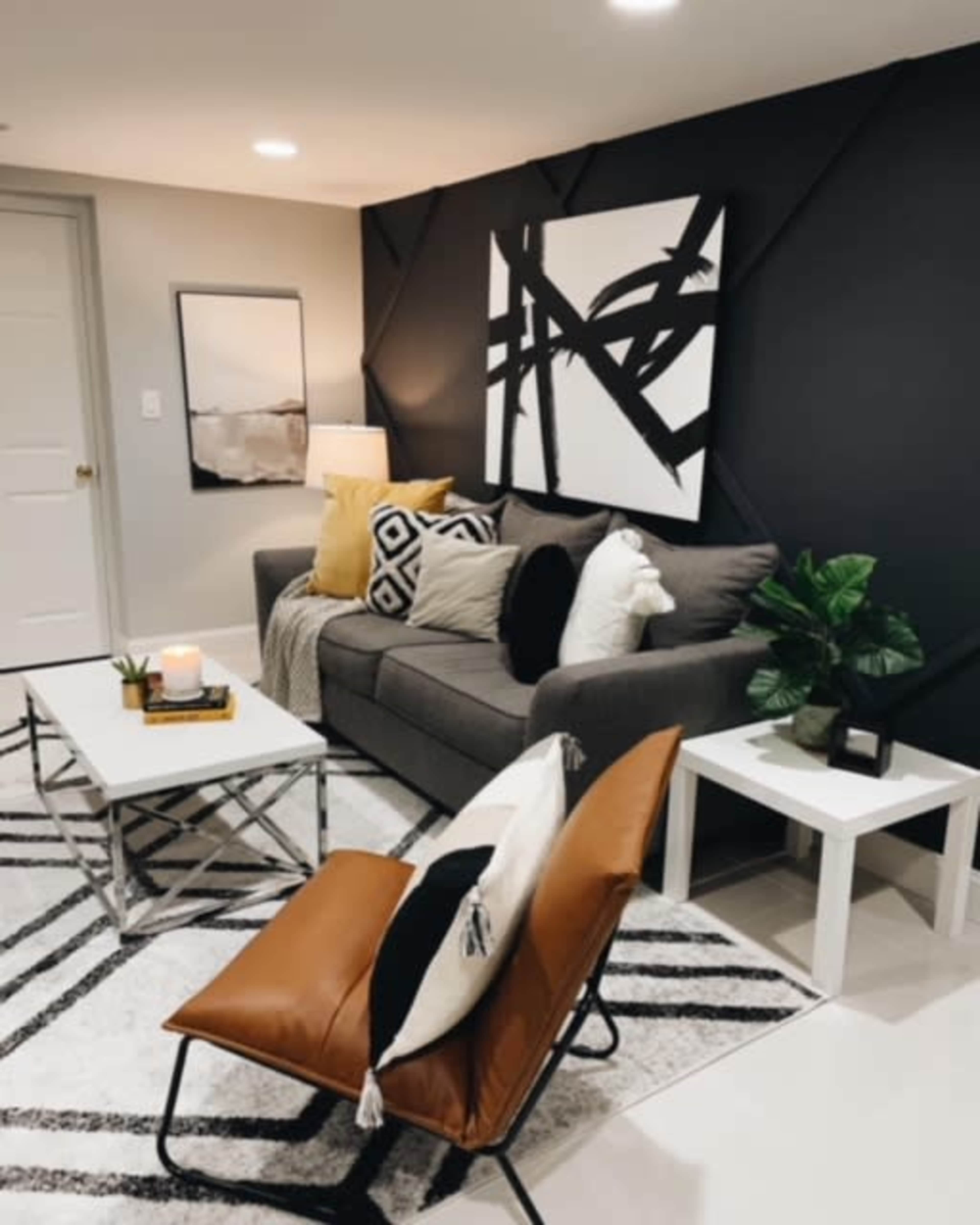 A modern living room featuring a gray sofa with decorative pillows, a coffee table, a patterned rug, and a white side table, all set against a dark accent wall with abstract art.