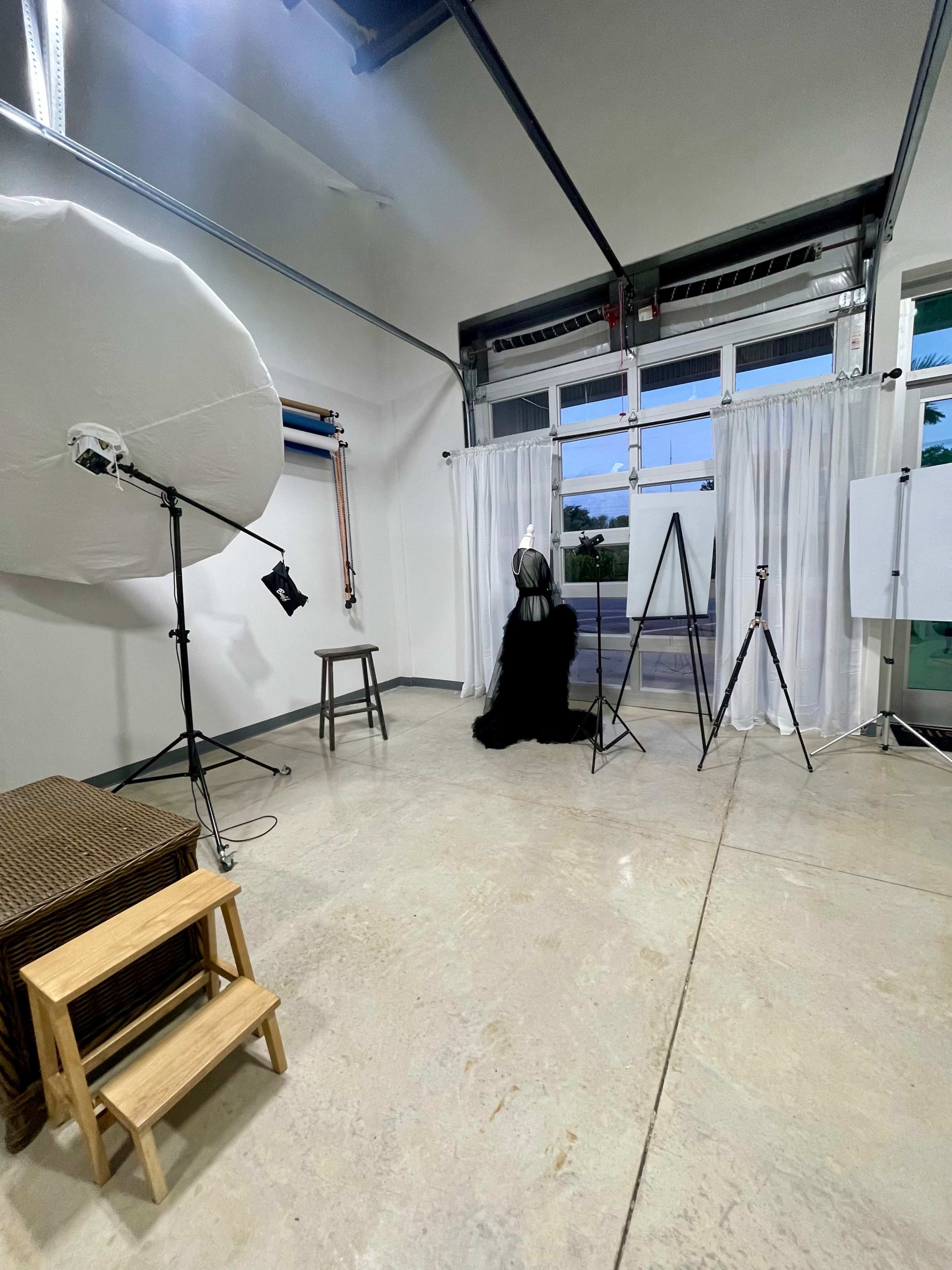 A photography studio setup with a backdrop, lighting equipment, and a model dressed in black sitting on a stool.