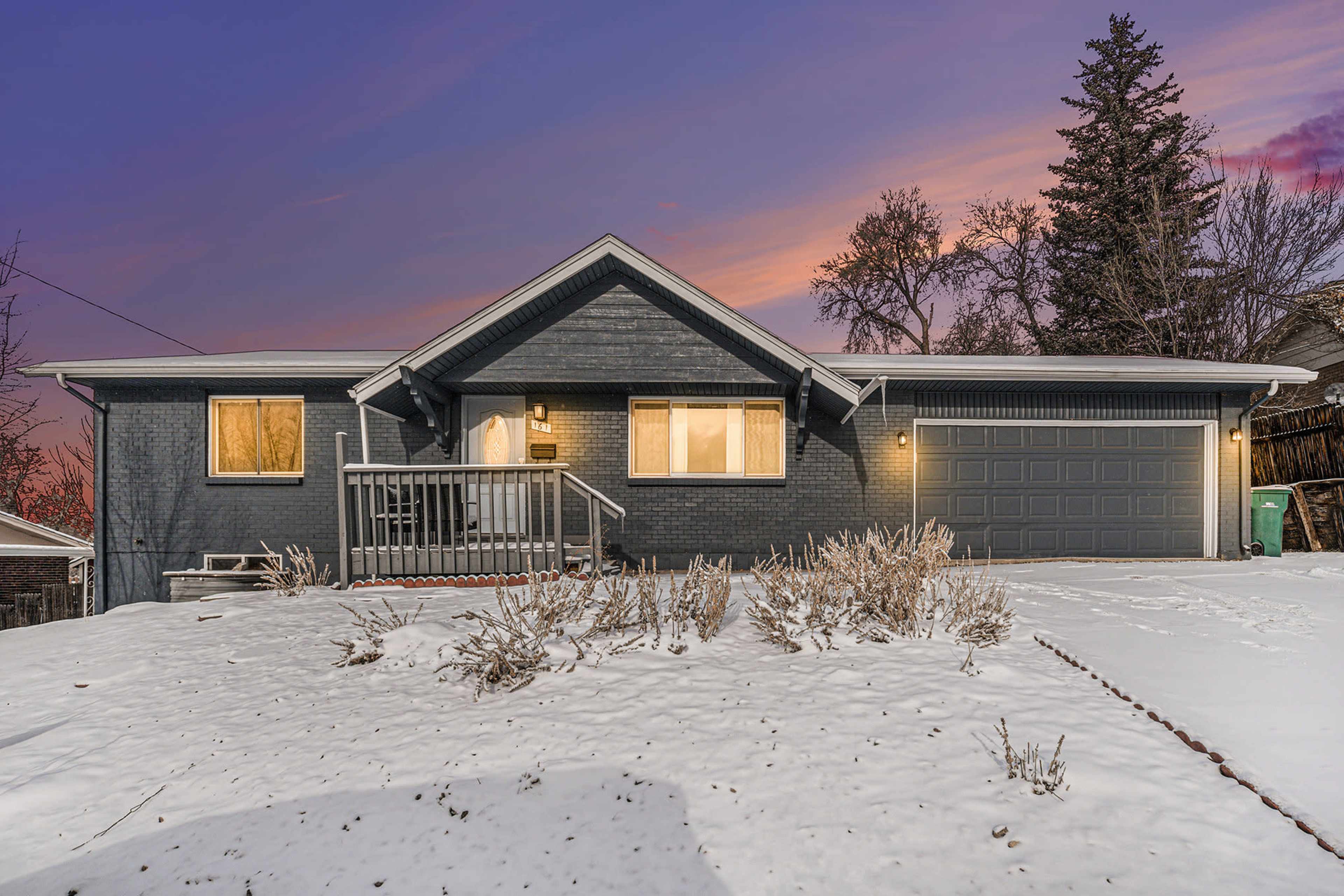 A single-story house with a gray exterior, a front porch, and a garage, surrounded by snow under a colorful sunset sky.