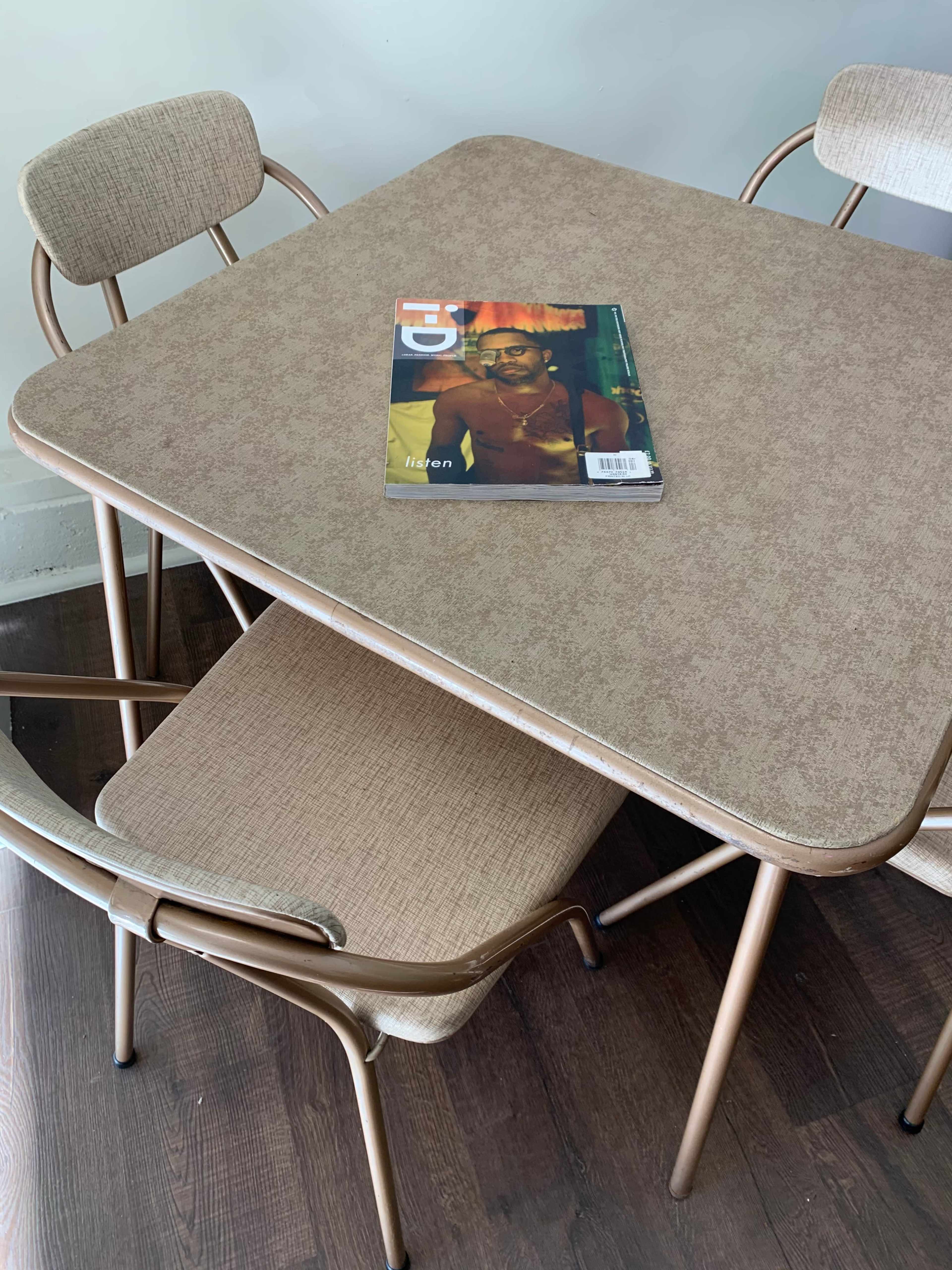 A square table with a magazine lying on top is surrounded by two chairs, one of which is stacked on the other.
