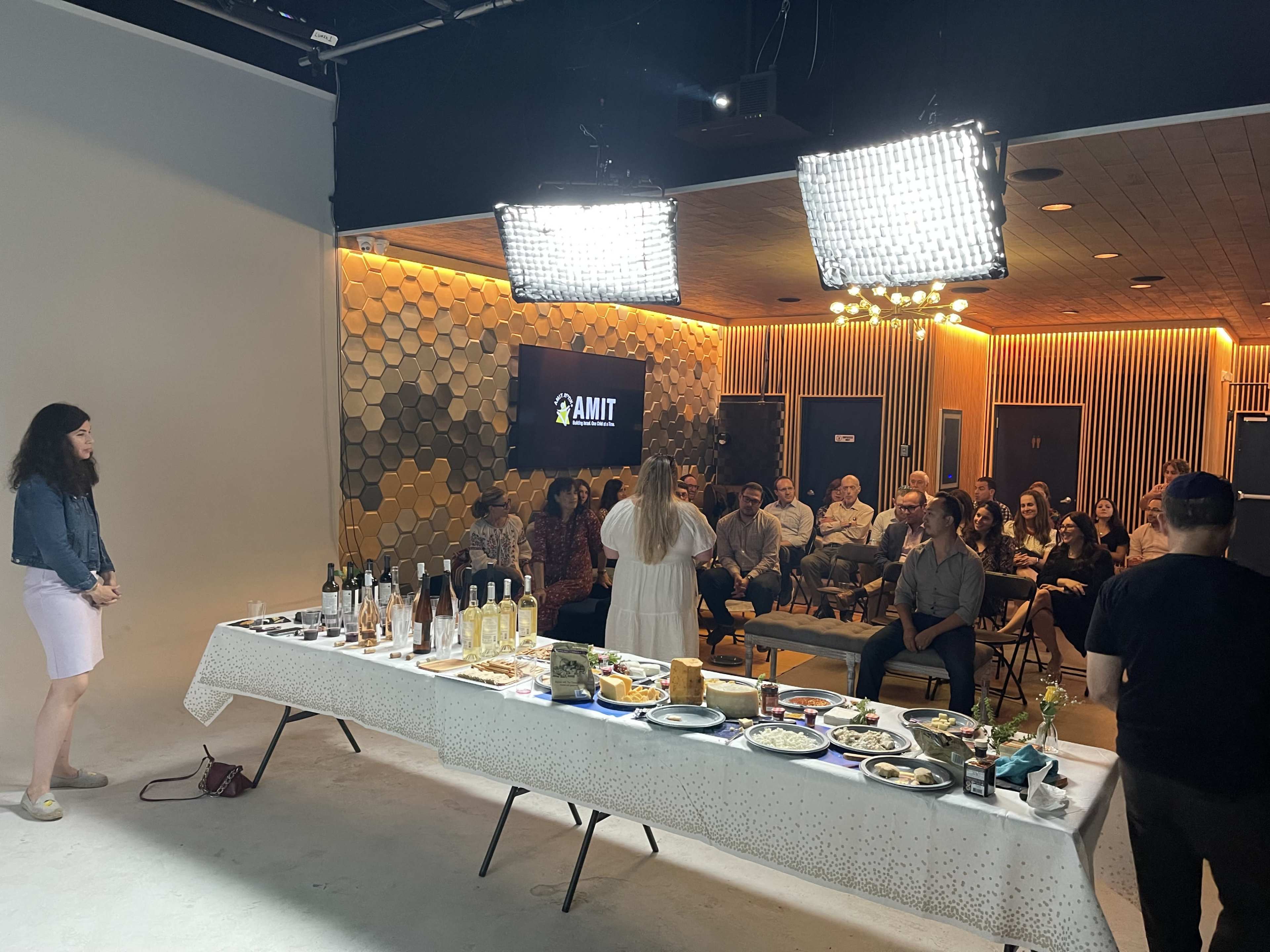 A group of people is seated in a well-lit studio, watching a presentation while a table adorned with various food and drink items is set in the foreground.