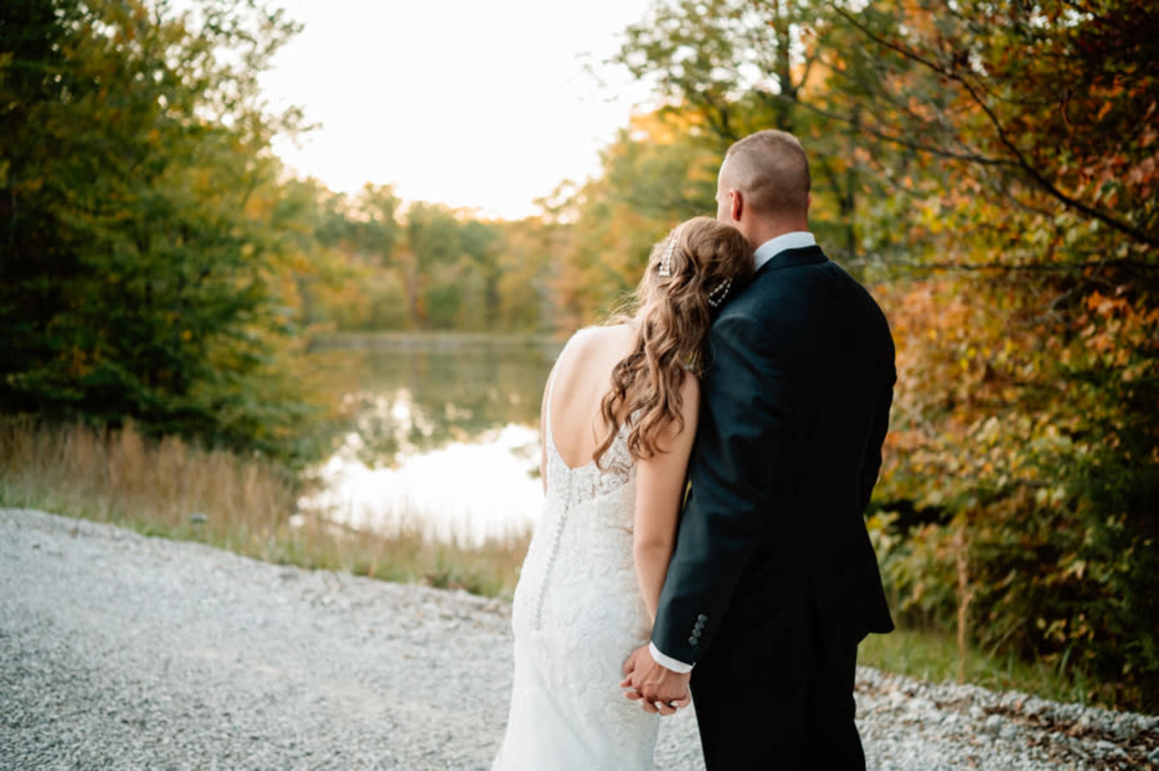 A couple holds hands while facing a serene lake surrounded by autumn foliage.