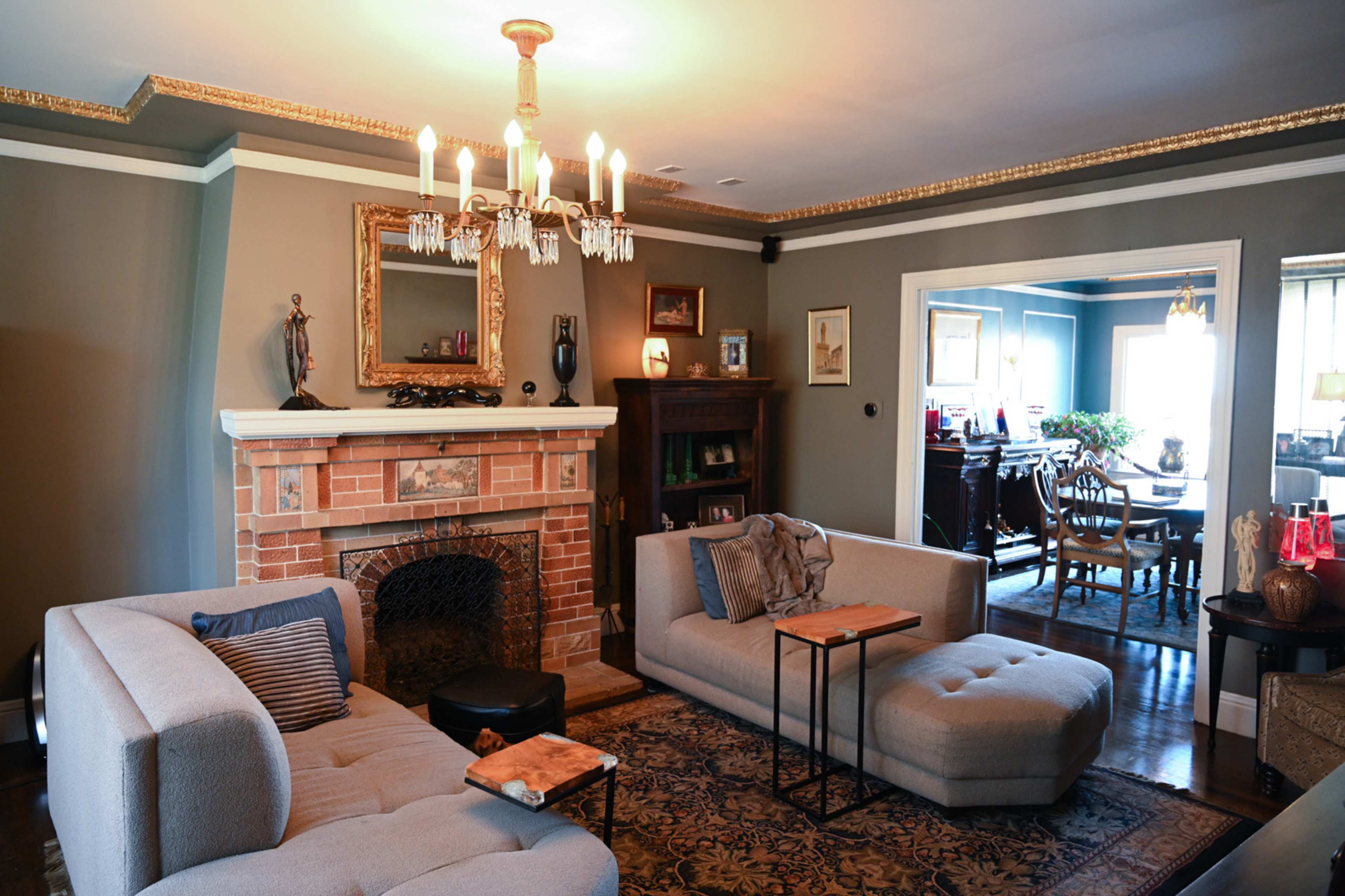 The living room features a brick fireplace with a decorative mirror above it, surrounded by two sofas and a coffee table, with a dining area visible through an adjoining door.
