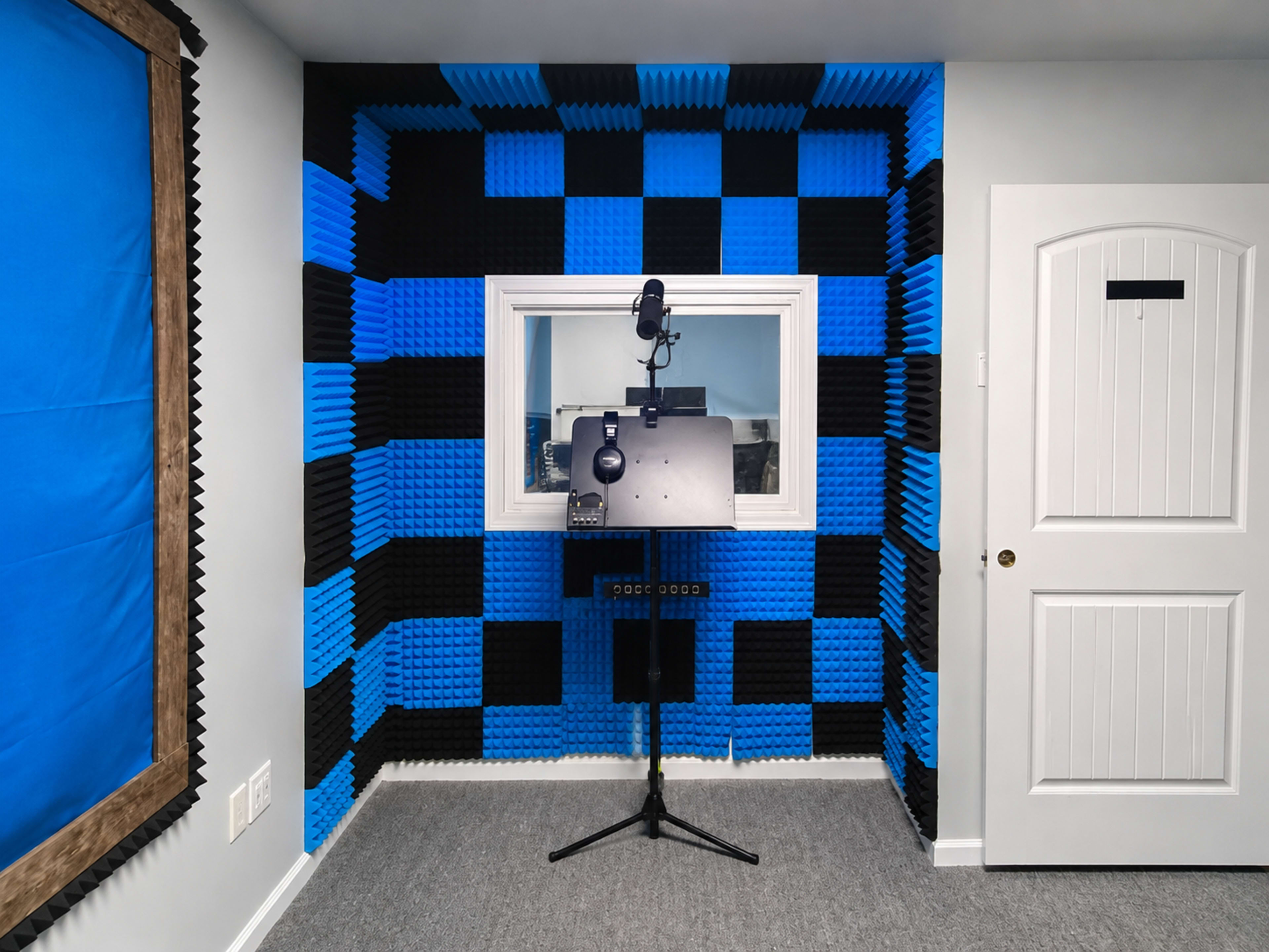 The image shows a soundproof recording studio featuring blue and black foam wall panels, a microphone setup on a stand, and an observation window.