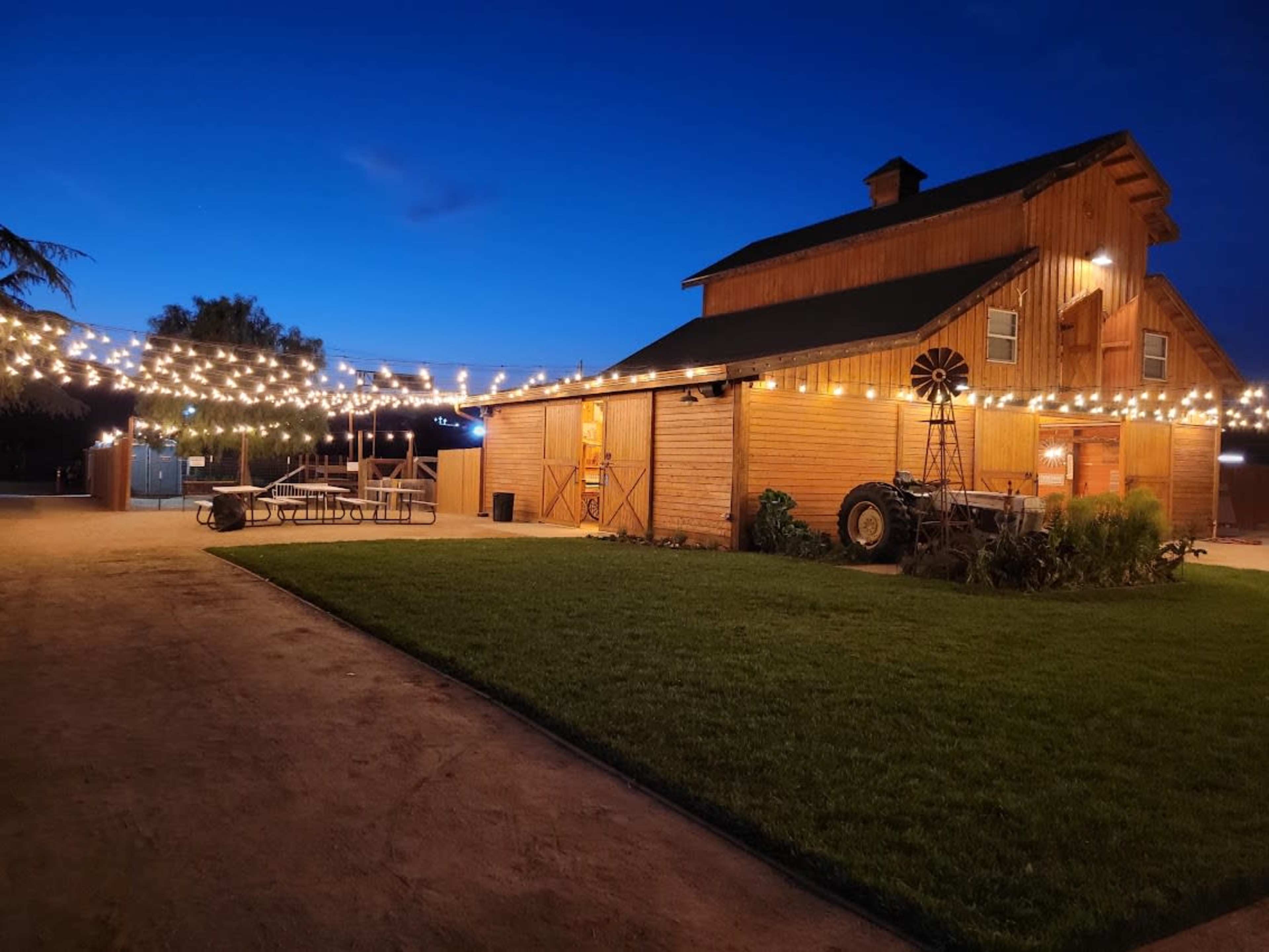 A wooden barn is illuminated by string lights under a twilight sky, with a grassy area and picnic tables nearby.