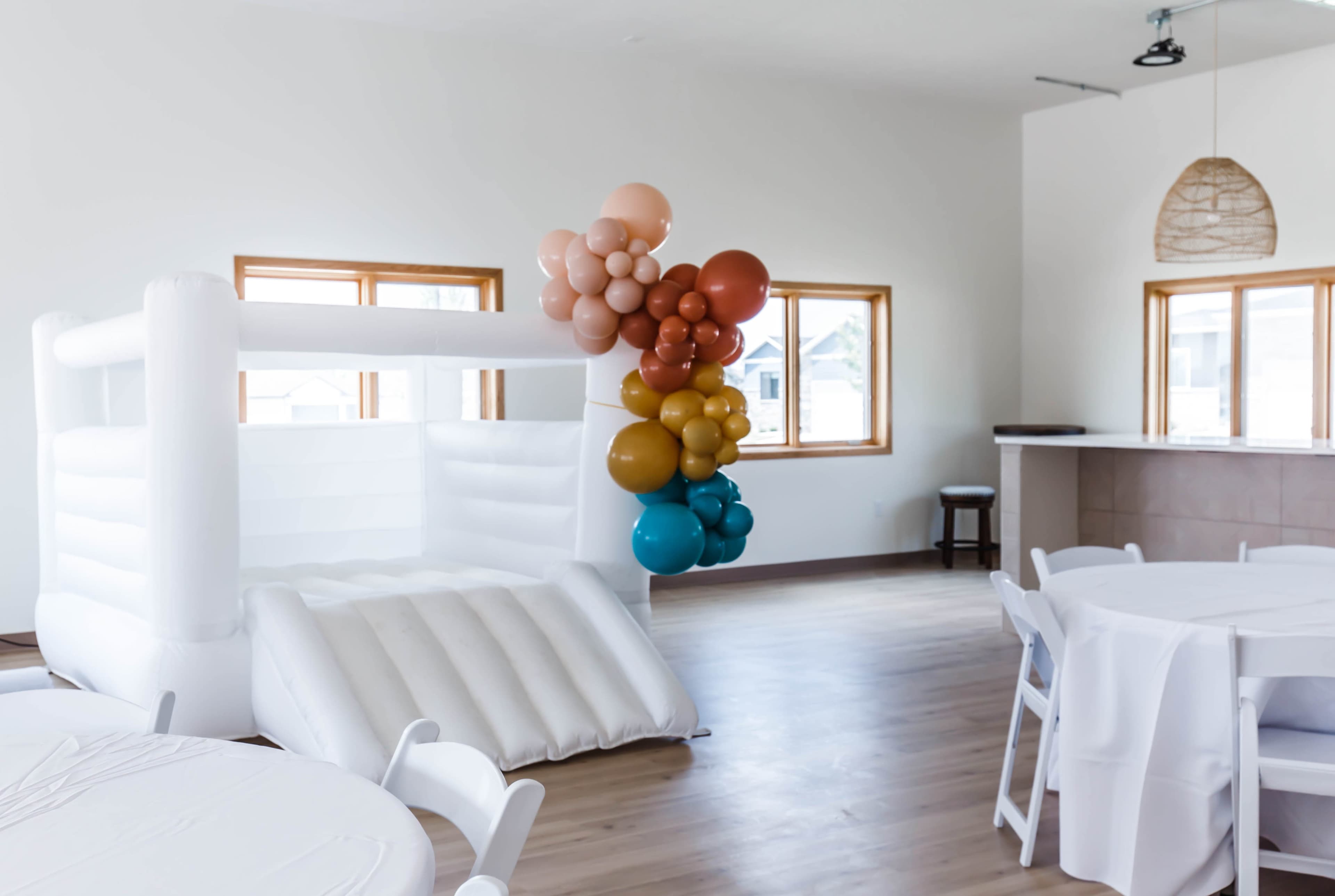 A spacious indoor party area featuring a white inflatable bounce house and a colorful balloon arrangement near the windows.