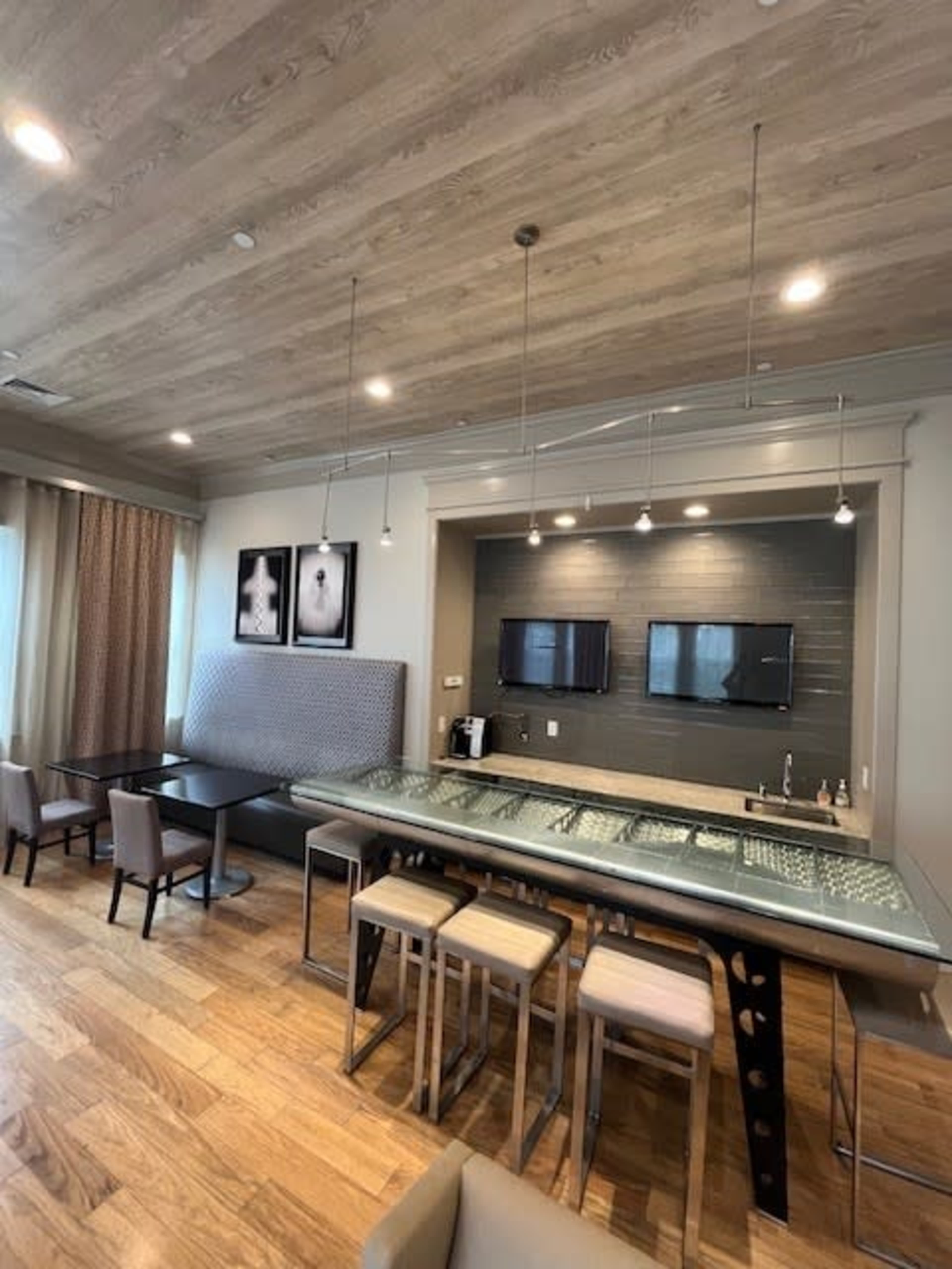A modern kitchen and dining area features a glass-topped bar with metal legs, two flat-screen TVs mounted on the wall, and wooden flooring accented by light fixtures.