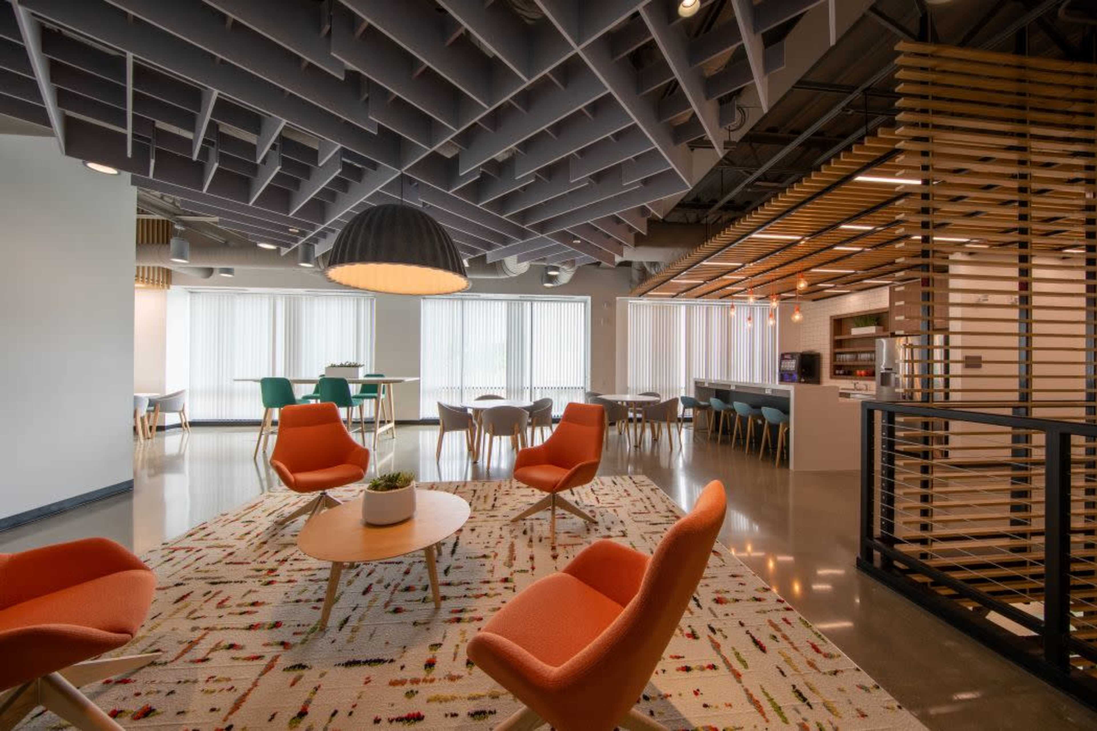 The image shows a modern office lounge area with orange chairs, a round wooden table, and large windows letting in natural light, complemented by a patterned rug and a kitchenette in the background.