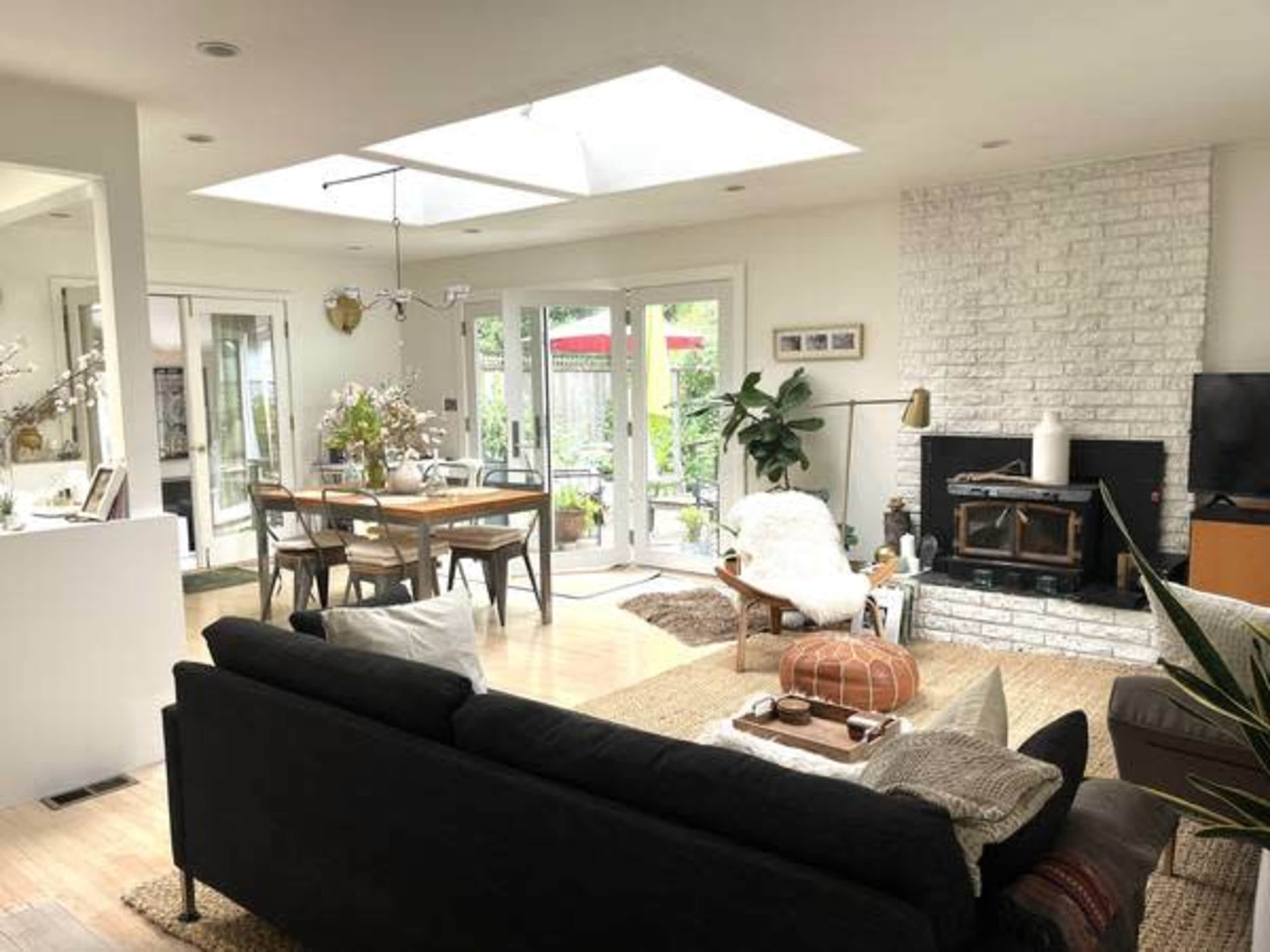 A modern living room features a black sofa, a coffee table, and a white brick fireplace, with a dining area and large windows providing natural light.